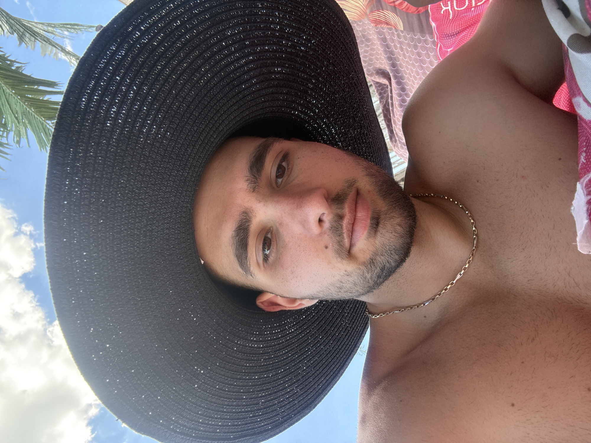A young man with dark hair and a beard is shown from the chest up, wearing a large black hat that casts a shadow over his face. He is wearing a gold chain around his neck and appears to be lying down outdoors, with a bright blue sky and palm fronds visible in the background. The man is on what appears to be a beach or poolside, with patterned fabric visible beneath him.