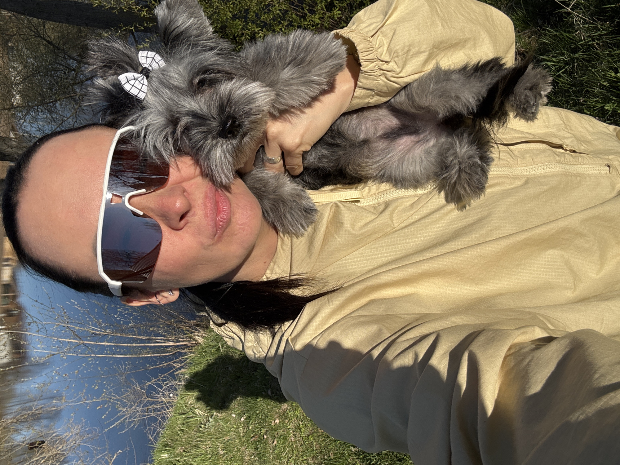 A woman wearing white framed sunglasses and a tan jacket is holding a gray Schnauzer dog with a checkered bow in its hair, belly facing the camera. The woman has dark hair and a silver ring on her finger. The background shows a body of water with reflections of trees and sky, and green grass. This location may be in the city of Chaikovsky, Russia.