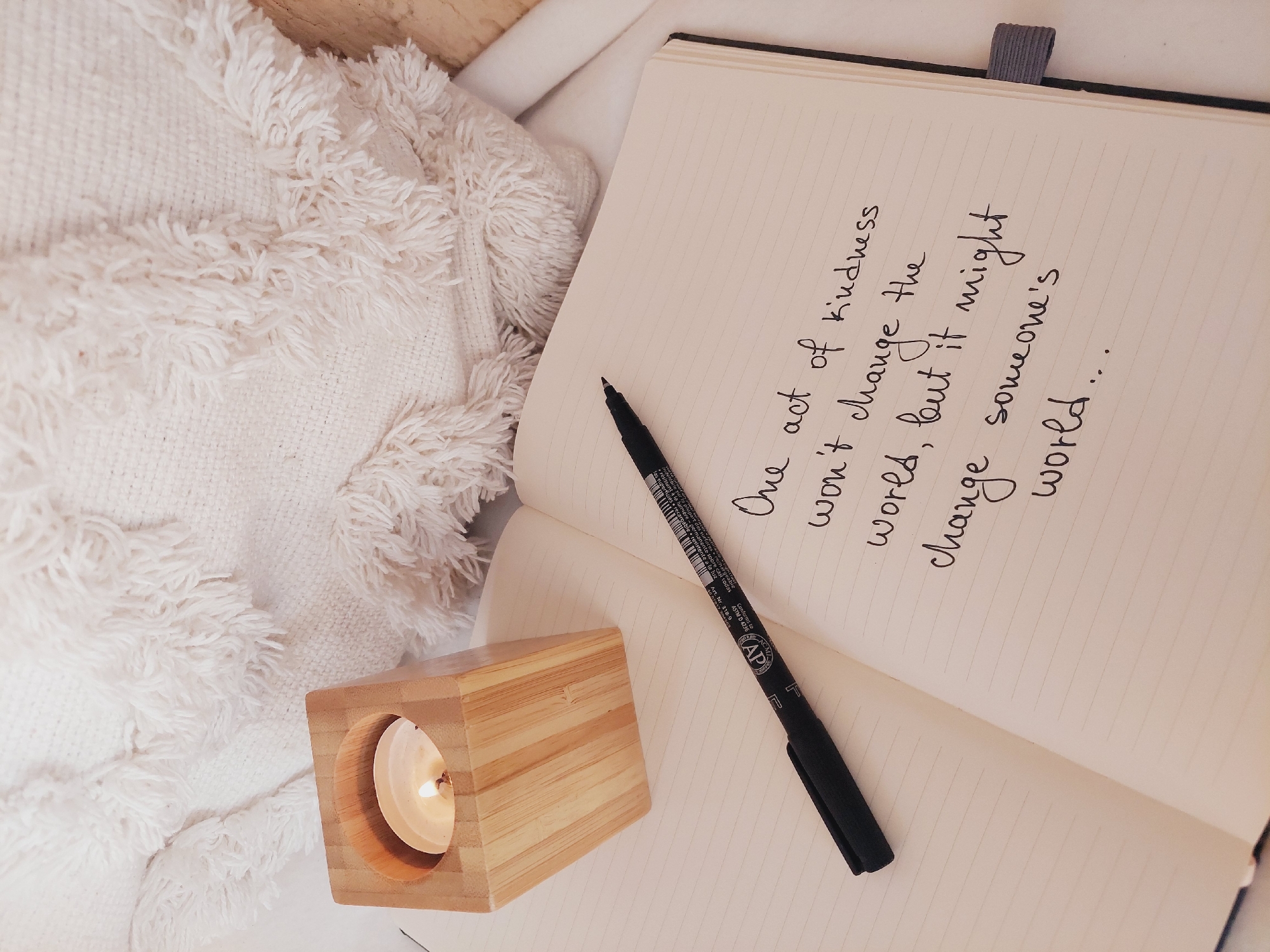 The image shows a notebook open to a page with handwritten text. A black pen rests on the notebook. Beside the notebook is a wooden candle holder with a lit candle inside. In the background, a white, textured textile is visible. The scene appears to be indoors, with soft lighting. There is no indication of the city of Sofia, Bulgaria.