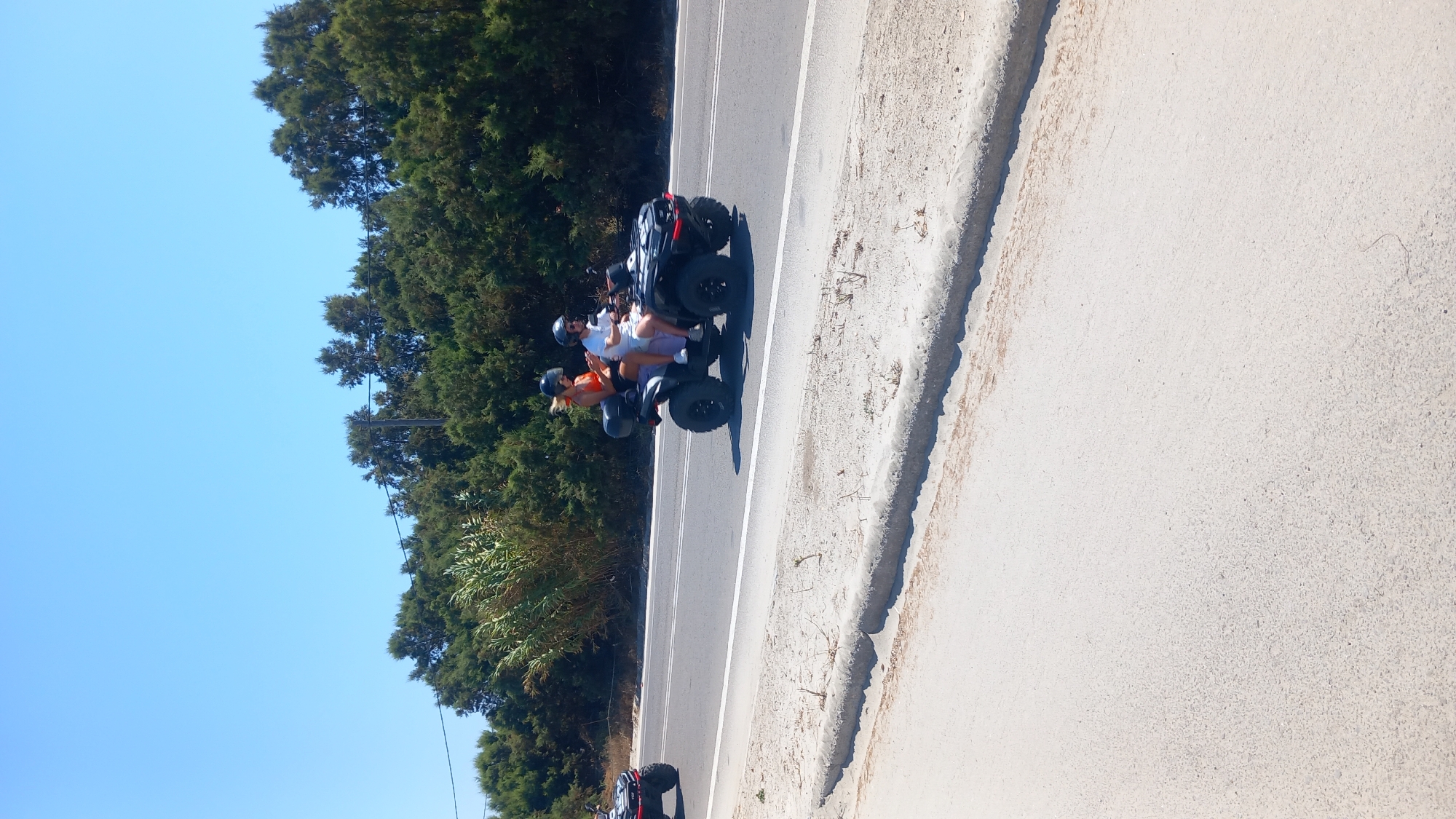 Two people are riding on a black ATV on a paved road. The ATV has two seats, and both individuals are wearing helmets. The person in the front is driving, and the person behind them is a passenger. They are both wearing casual clothing. The ATV is black and appears to be a four-wheeler with large tires. Behind the ATV, a line of green trees and foliage forms a dense natural border along the side of the road. The sky is clear and blue, suggesting a sunny day. A sandy area is visible on the left side of the road, bordering the pavement. Another red ATV is partially visible in the distance on the left side of the image.