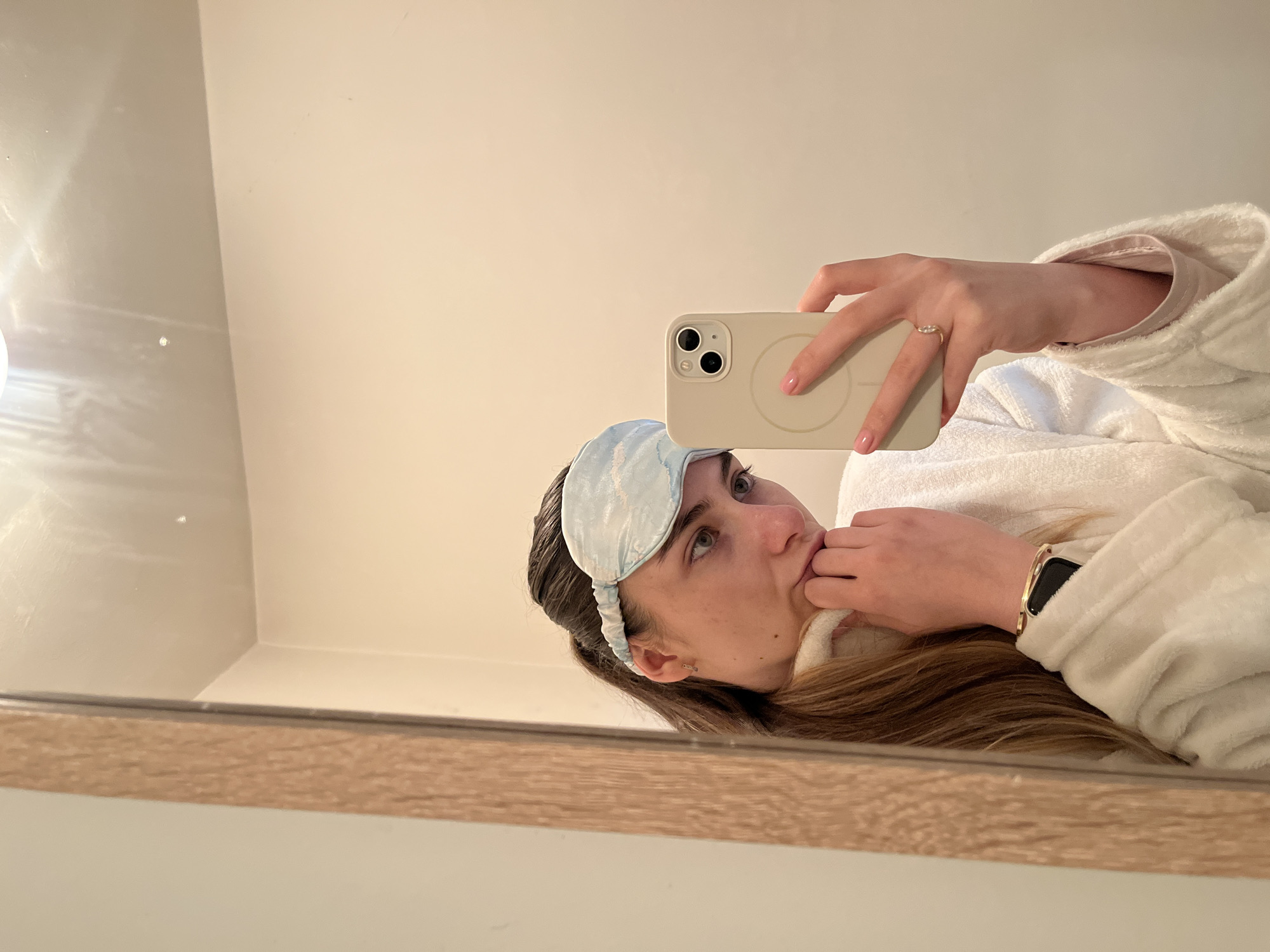 A woman is reflected in a mirror, holding a smartphone in her right hand to take a selfie. She is wearing a light blue sleep mask over her forehead, a white robe over a light pink shirt, and a gold wristwatch on her left wrist. She has brown hair and is looking toward the upper right of the frame, with her left hand near her mouth. The smartphone is light beige with a circular MagSafe charging interface on the back. The background consists of a neutral-colored wall, and the mirror has a light brown frame. The location appears to be indoors, likely a bathroom. The city is Unknown, Unknown.