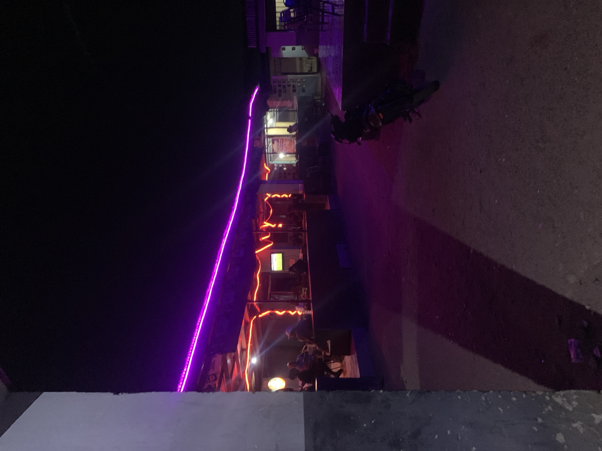 The image shows a dimly lit scene, seemingly at night, in the vicinity of New Stadium, Nigeria. A building is adorned with both purple and red neon lights. People are seated inside, under the warm glow of interior lighting and the red neon accents. Outside, a motorcycle is parked. The ground is paved. A low wall is visible in the foreground.