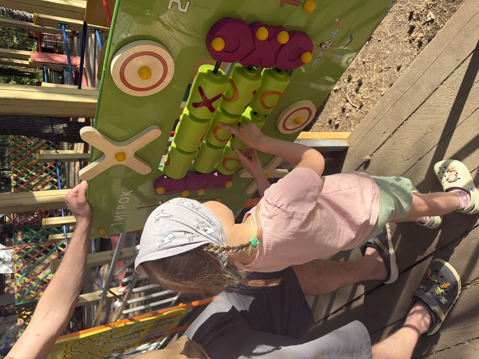 A young girl is interacting with a green, purple, and yellow play structure with rotating cylinders, as an adult steadies it; the structure appears to be part of a playground. The girl is wearing a light pink shirt, green shorts, a patterned headscarf, and white sandals. The adult is wearing dark shorts and dark sandals. The setting appears to be an outdoor playground, possibly in a park, located on a wooden platform or deck. The location is identified as the city of Chaikovsky, Russia, based on the Cyrillic text "Сделано в России" (Made in Russia) on the play structure, though the specific location within Chaikovsky cannot be determined from the image.