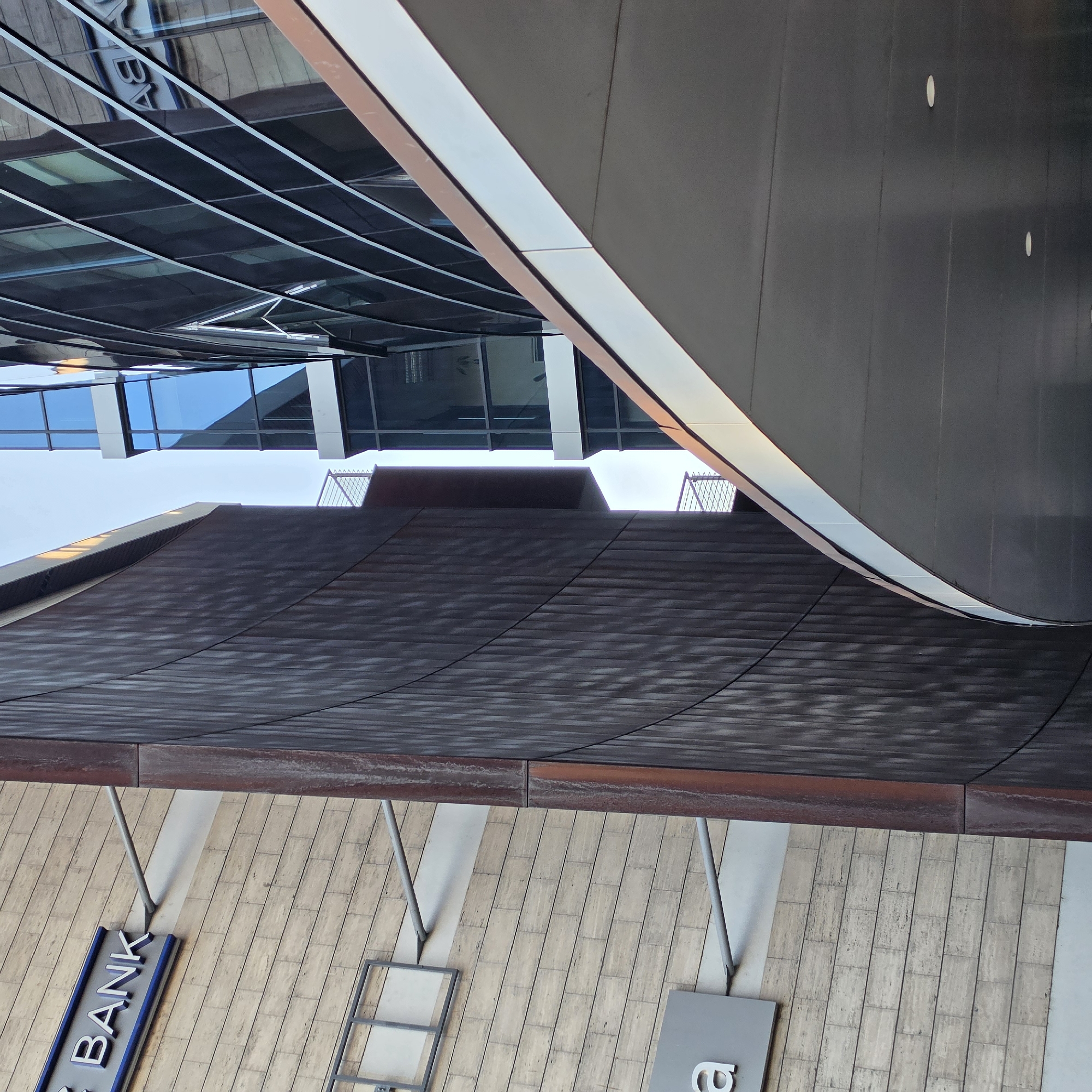 The image depicts an upward-angled view of the exterior of a building, likely in an urban setting. The foreground shows a section of a building facade with a horizontal paneling design, possibly made of stone or similar material. A sign with the word "BANK" in blue letters is visible on the lower left. Above, there's a dark, curved, horizontal structure that appears to be an overhang or architectural feature with a textured surface. Above this structure, the reflection of the same bank is seen in another building. The upper portion of the image features part of a modern glass and steel building, suggesting a contemporary architectural style. The overall impression is of a city building exterior, emphasizing a blend of architectural styles. The Podgorica, Montenegro, location isn't visibly supported by the image.