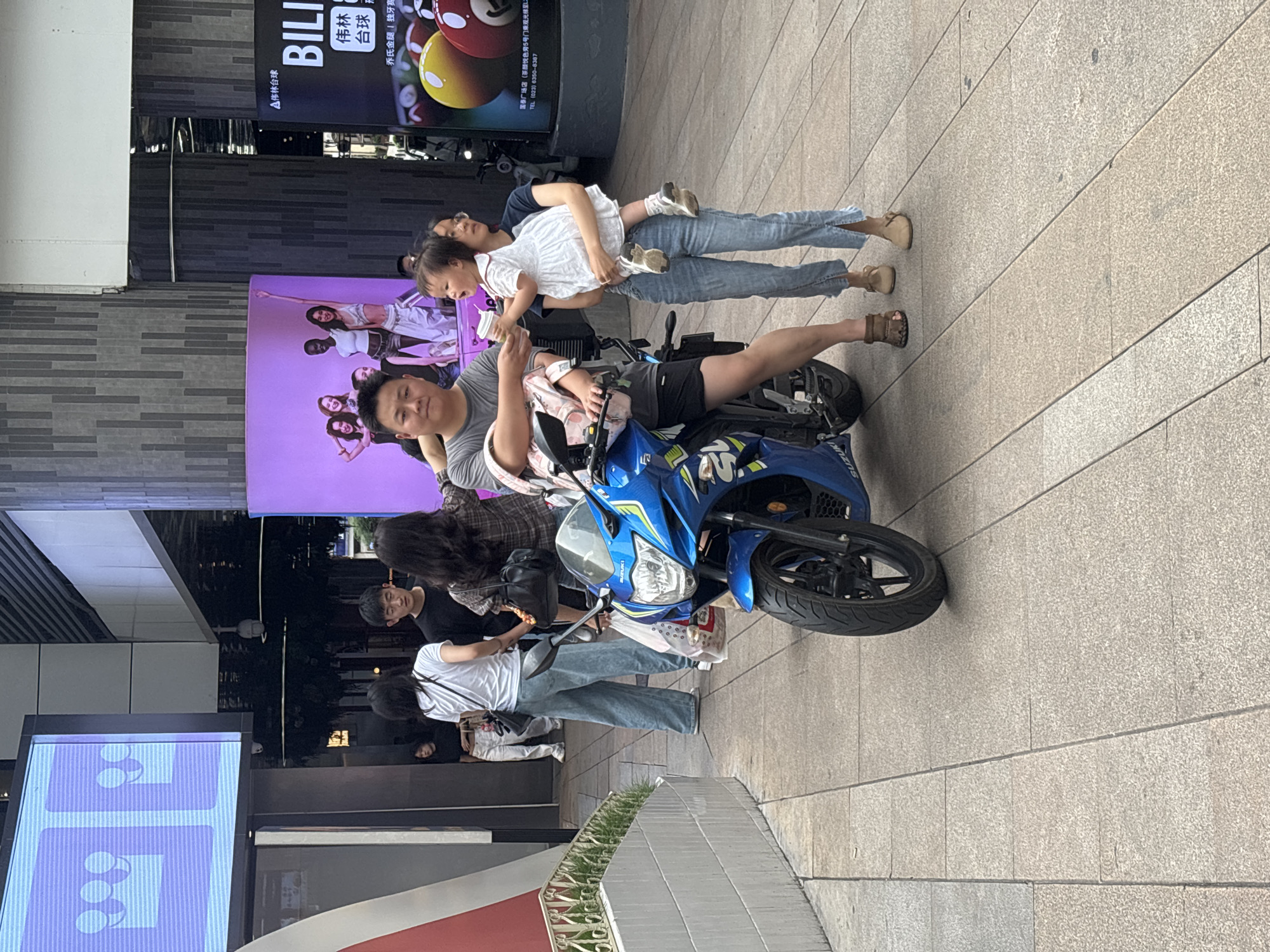 The image shows a street scene in Yuzhong District, China, featuring a man sitting on a blue Suzuki motorcycle. He is wearing a grey shirt and black shorts. A woman stands behind him, holding a young child. They are standing on a paved walkway in front of shops with digital billboards. The background includes advertisements for billiards (BILI 伟林 台球). There are other pedestrians nearby.