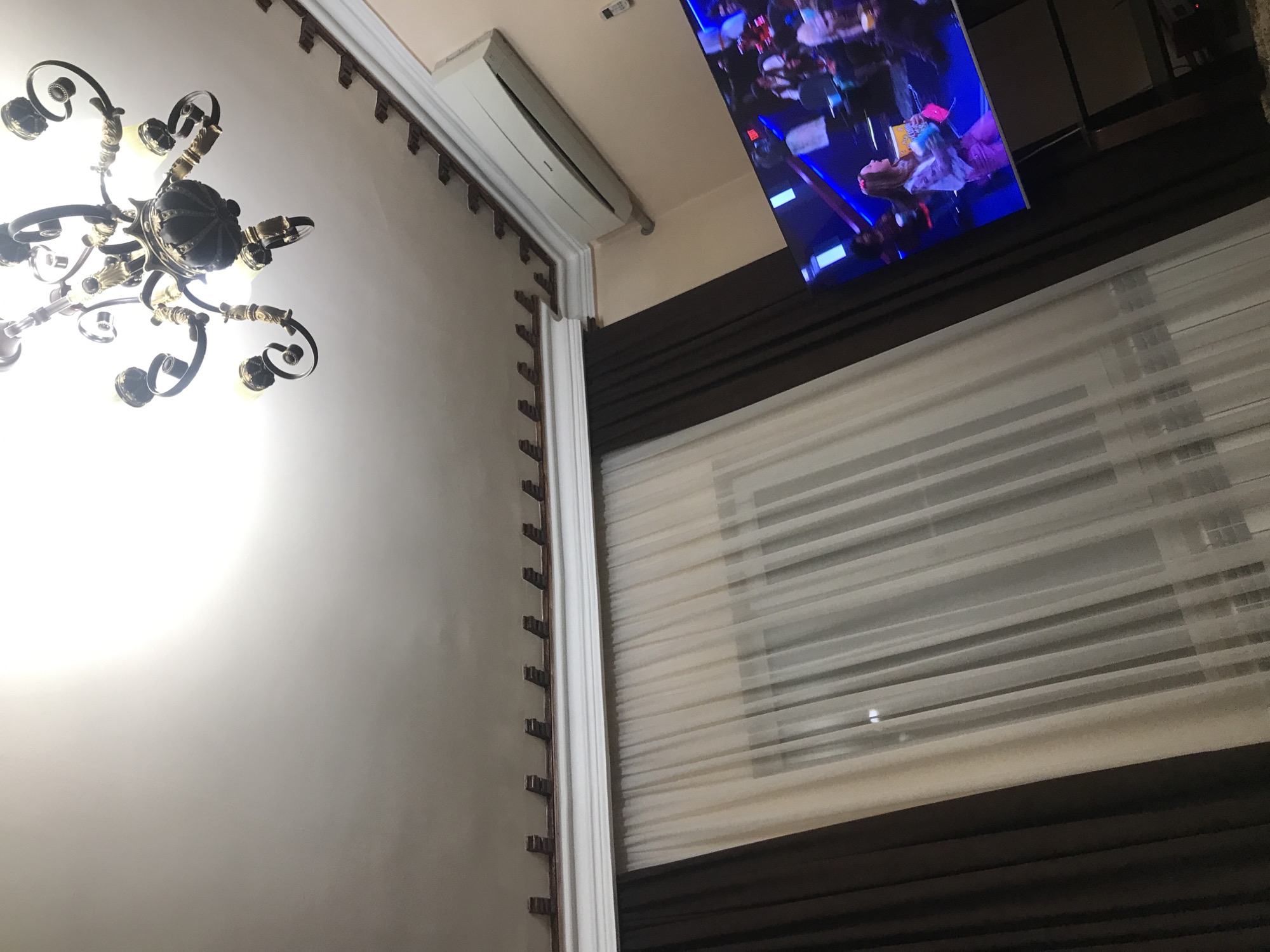 The image captures an indoor scene, likely in a home in Yerevan, Armenia, based on the architecture and decor. A chandelier is visible in the upper-left corner, hanging from the ceiling. Part of an air conditioning unit is mounted near the ceiling. A television displays an event, possibly a concert or awards show, featuring people on stage. A window with closed blinds is present. The walls are painted in a light color, and ornate trim decorates the edges.