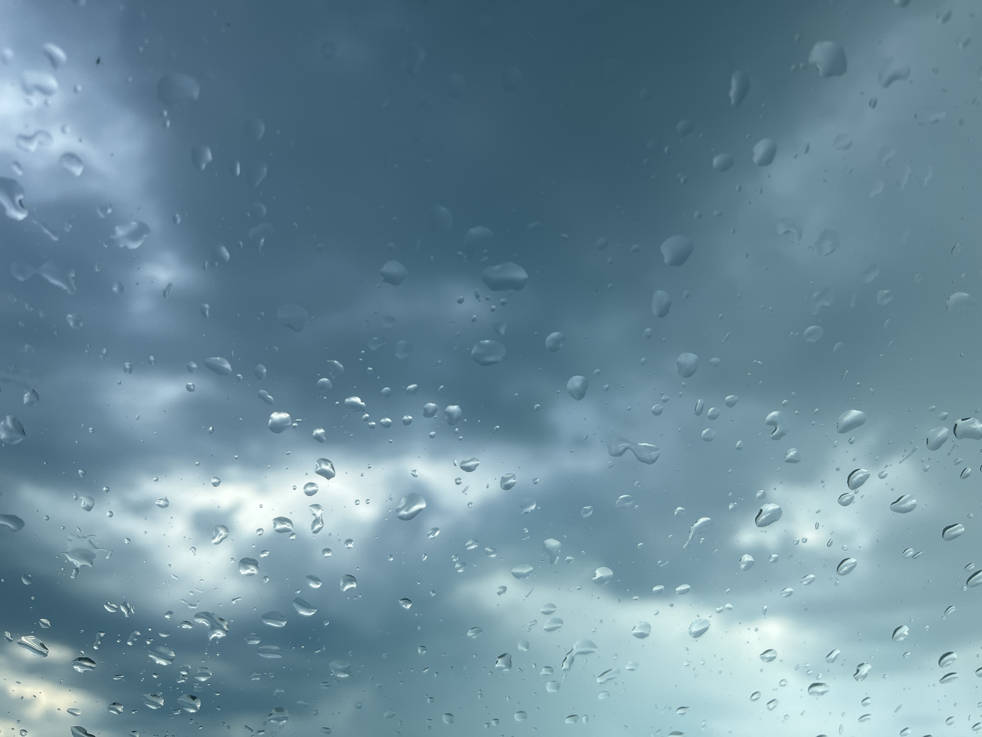 The image shows a view through a wet surface, possibly a window, with raindrops scattered across the glass. The background is a cloudy sky, with varying shades of gray and patches of light breaking through the clouds. No people, subjects, or specific objects are visible beyond the raindrops and the sky. No actions or interactions are depicted. The location in Varna, Bulgaria is not specified.