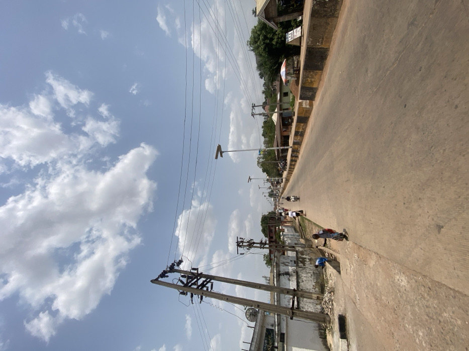 This image depicts a street scene in Afikpo, Nigeria, under a bright, partly cloudy sky. The perspective is from a low angle, looking down a two-lane, paved road that extends into the distance with a gentle incline.
In the left foreground, a tall, leaning wooden utility pole with multiple cross arms and numerous power lines dominates. Below it, two children are present on the unpaved shoulder. One child, wearing a blue top, is stooping near a concrete drain channel. The second child, in a light shirt and dark shorts, stands upright, facing the street while holding a red object, possibly a backpack. Behind the utility pole and children, a white building with a satellite dish on its roof is visible, fronted by a low concrete wall.
The paved road occupies the central and right portions of the frame. On the right side, a low concrete barrier, painted in alternating black and yellow/tan segments, separates the road from the adjacent area. A line of modern streetlights, each topped with a solar panel, runs alongside this barrier into the distance. Further down the road, several pedestrians walk on the left side, and a motorcycle carrying at least one individual travels away from the viewer.
On the right side of the road in the midground, various single-story structures or shops line the street. A white and red striped umbrella provides shade in front of one of these establishments, near a white sign displaying text, including "L.P.G." and "STATION." Green foliage from trees and bushes is visible behind these structures. The scene is illuminated by strong daylight, casting minimal shadows directly beneath objects.