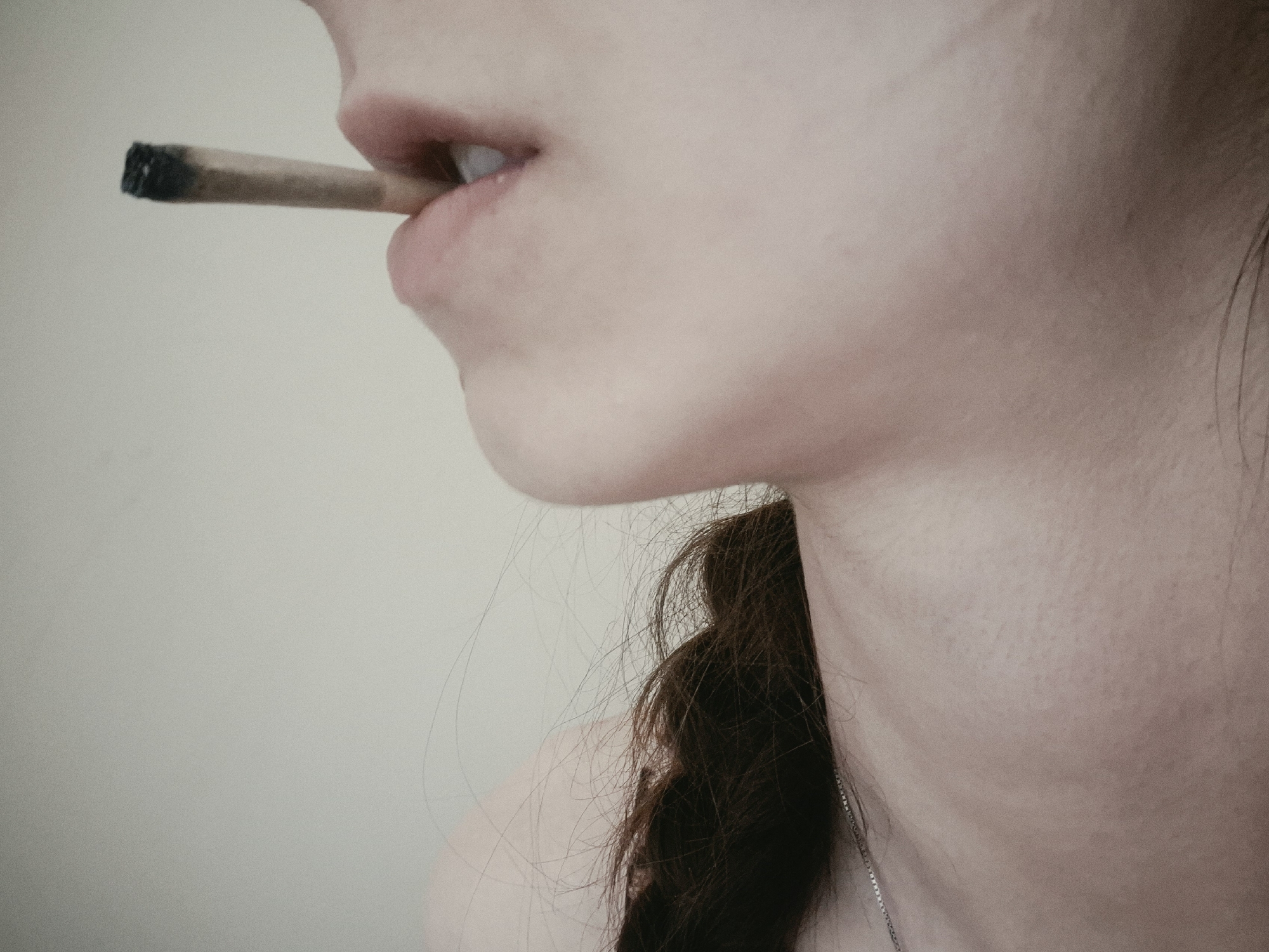 The image shows a close-up of a person's mouth and neck. The person is holding a lit cigarette between their lips. The background is a plain, light-colored wall. There is no indication of the location in the city of Sofia, Bulgaria.