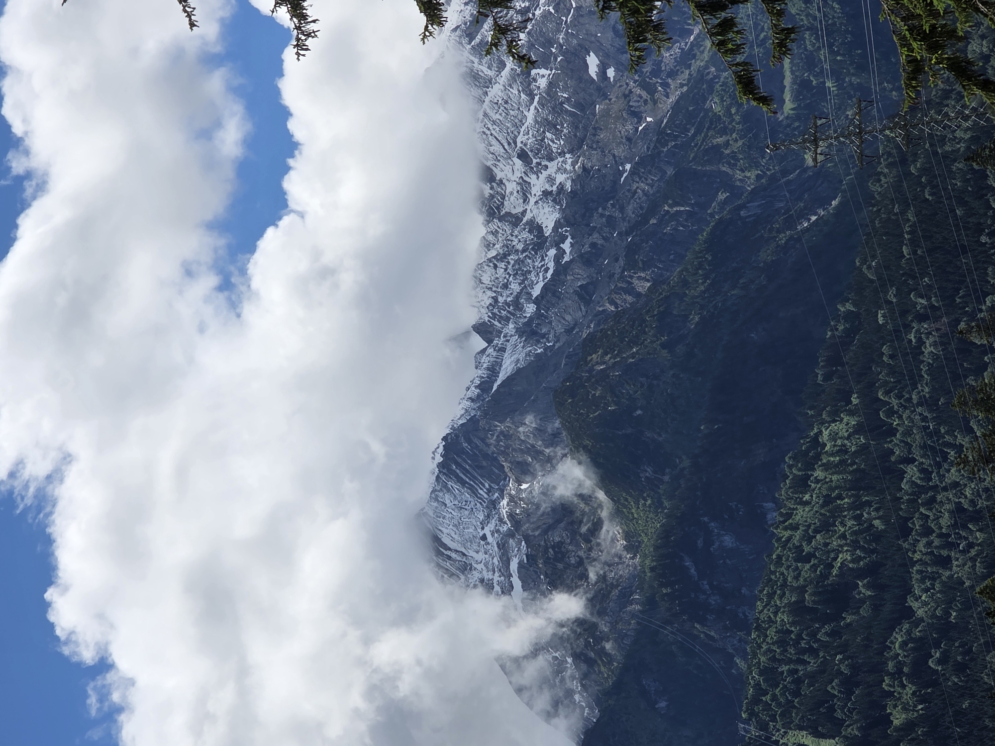 A dramatic mountain range dominates the frame, its peaks partially obscured by thick, white clouds. The higher elevations are dusted with snow, contrasting sharply with the dark, rugged rock faces. Lower down, steep slopes are blanketed in dense, dark green coniferous forests. In the foreground, a power pylon stands tall, supported by wires that stretch across the scene, hinting at human infrastructure in this wild landscape. The sky above is a vibrant blue, with patches of white clouds adding texture and depth. The lighting suggests it is daytime, possibly late morning or early afternoon. The location is identified as Unknown, Unknown.