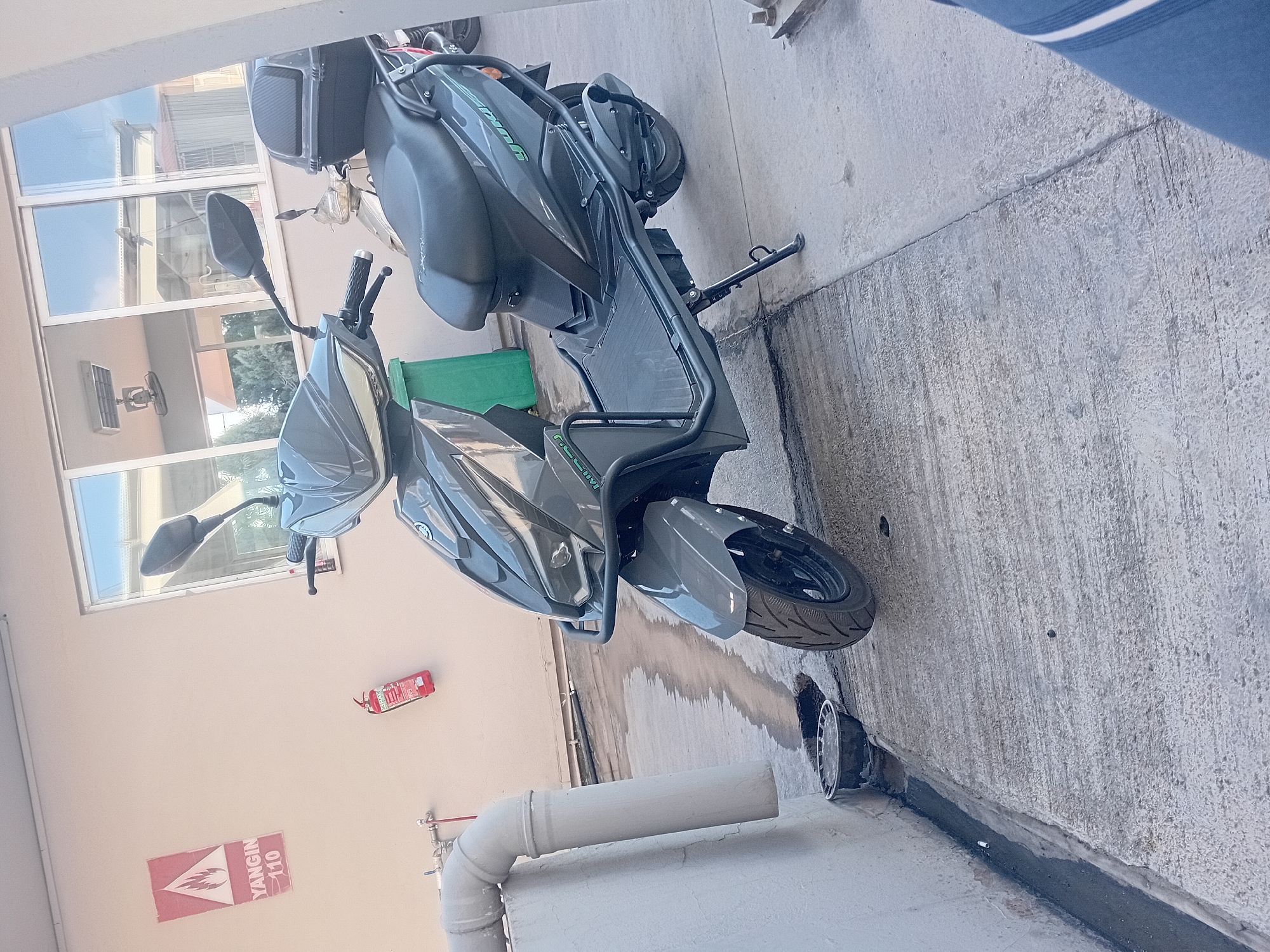 In the city of Onikişubat, Turkey, a grey scooter is parked on a concrete surface next to a building. The scooter has a black seat and a black storage box on the rear. A fire extinguisher is mounted on the wall of the building, and a red and white sign with flames and "YANGIN 110" is also visible on the wall. A green trash can is next to the scooter. The ground is wet in places, suggesting recent rain or cleaning. The environment appears to be an outdoor commercial or industrial area, with windows visible in the background.