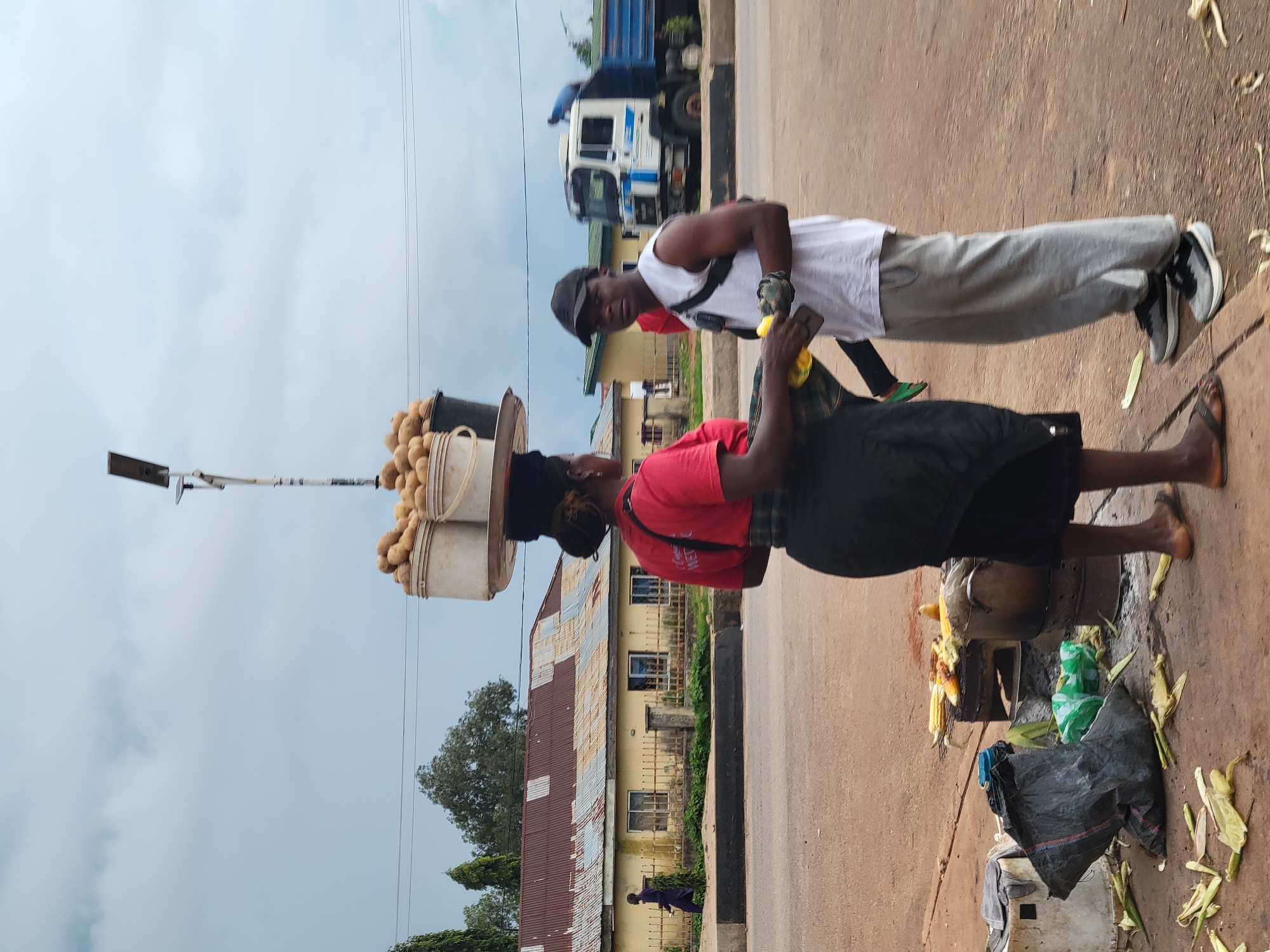 In Gura Top, Nigeria, a street vendor, a woman wearing a red t-shirt and a black skirt, balances a large tray of potatoes on her head. She is interacting with a man wearing a white sleeveless shirt, grey pants, and a black baseball cap. He is holding a yellow object, possibly an ear of corn, and a mobile phone. The scene takes place on a dusty road with buildings in the background. To the left, a man in a purple garment walks past a yellow building with a corrugated metal roof. To the right, a large white and blue truck is parked. In the foreground, discarded corn husks and a large black plastic bag lie on the ground, indicating a food vending area. A metal grill with roasted corn and a large pot are visible near the woman's feet. The sky is overcast with grey clouds.