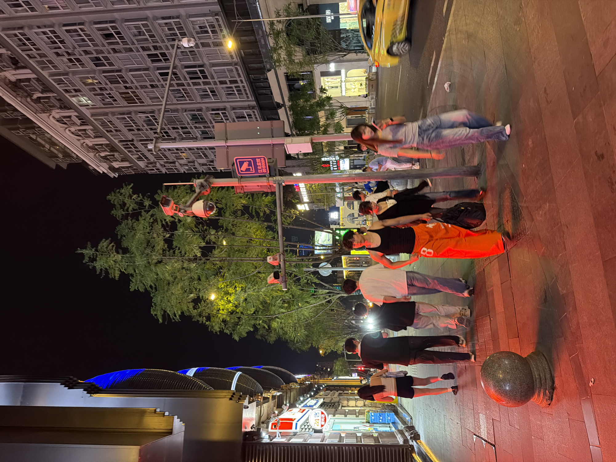 Captured at night, the scene presents a pedestrian street in the city of Liangjiang, China. Pedestrians of various ages and attires are observed strolling on the pavement, some wearing face masks. Overhead are security cameras, street lights, and trees. A building with a distinct architectural pattern is seen on the left, featuring blue decorative lighting. On the right, there's a road with a yellow taxi. Various establishments and advertisements are visible in the background, illuminated by bright lights. The pavement has red illumination.