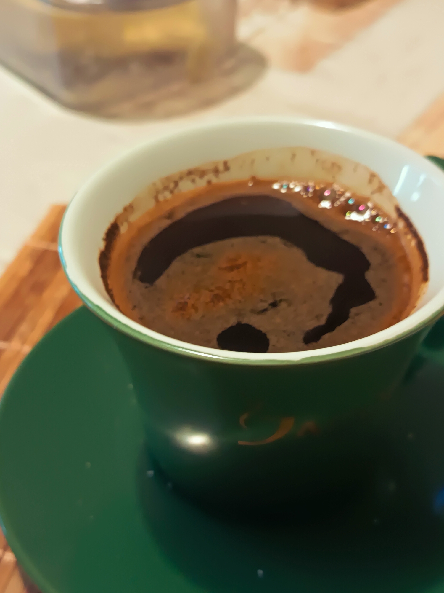 A close-up, slightly blurry shot shows a dark brown cup of coffee with a foamy surface, sitting on a green saucer. The cup has a white rim with a thin green outer edge. In the background, out of focus, is a clear glass jar with a light brown substance inside, possibly coffee grounds or sugar. The setting appears to be indoors, with a textured surface beneath the saucer and cup that could be a table. There are no people or any identifiable landmarks of Izmail, Ukraine visible in the image. The lighting suggests an indoor environment, possibly with artificial light contributing to the reflections on the coffee surface and saucer.