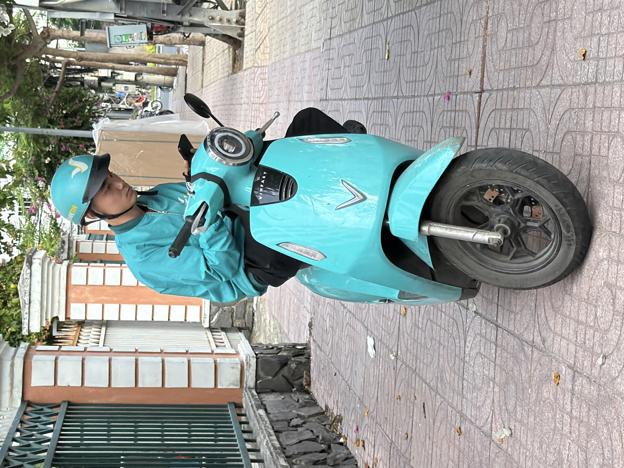 A young man wearing a teal jacket and helmet sits on a teal electric scooter on a tiled sidewalk in Nha Trang, Vietnam. He is looking down at his phone, which he holds in his left hand. The scooter has a VINFAST logo on the front. A metal fence is visible to the left of the man, and trees and buildings line the street behind him. Fallen leaves are scattered on the sidewalk.