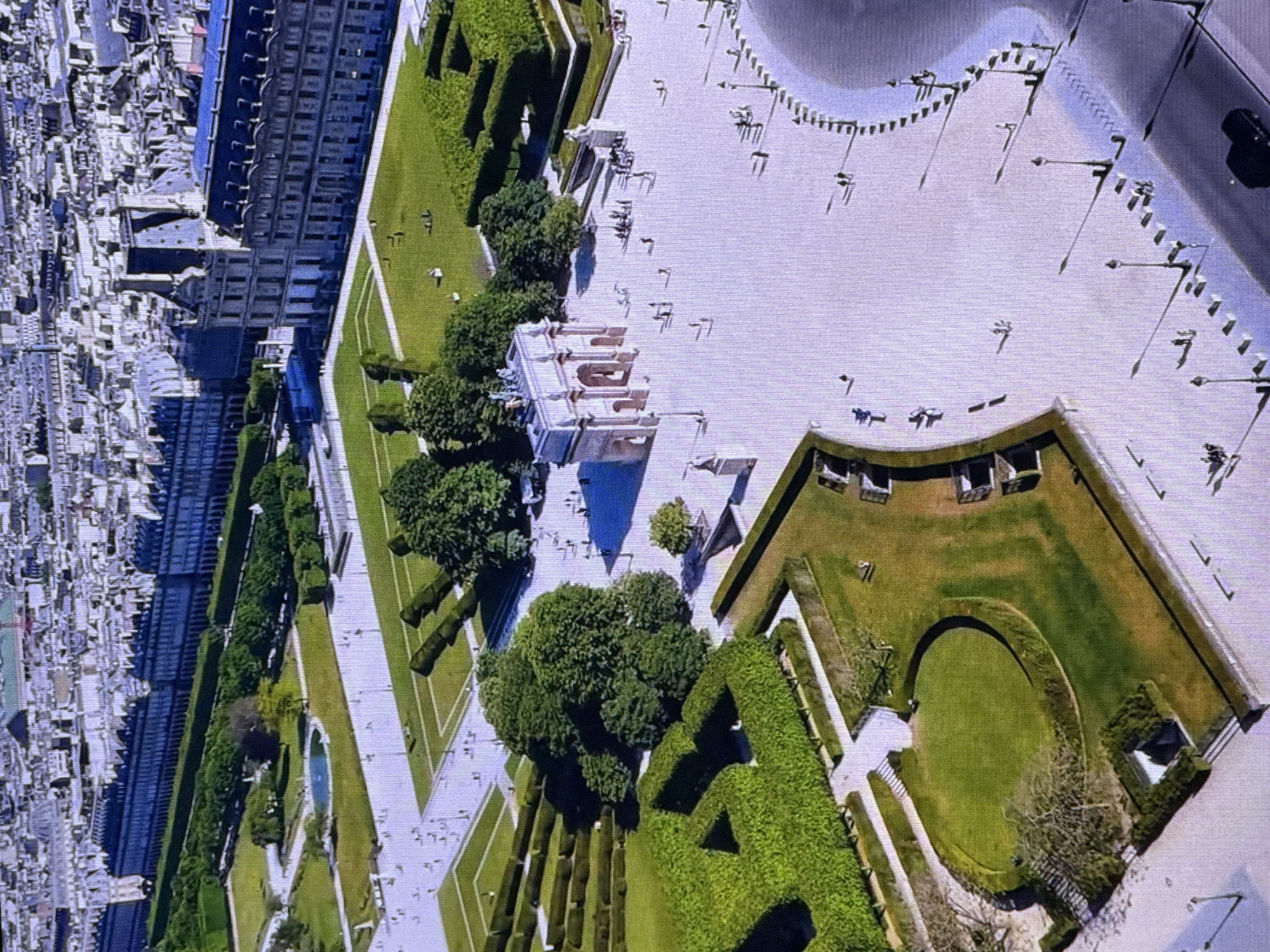 This aerial image captures a geometrically designed garden and courtyard area adjacent to a large, classical-style building and a dense urban landscape. The garden features manicured hedges, including a large, prominent hedge shaped into a letter, and a circular lawn bordered by a curved hedge. Pathways and paved areas are visible, with a small water feature or pond.
Scattered throughout the paved areas are numerous small figures, suggesting people are present, though their details are indistinct. There are also lamp posts with flags or banners, and a black vehicle is visible in the upper right quadrant of the image.
The scene appears to be an outdoor public space, likely a park or a garden area associated with a significant building, possibly a museum or historical landmark given the architectural style of the building in the background. The lighting suggests it is daytime, and the weather appears clear.
The image's orientation is such that the urban landscape on the left is vertical, with the garden and courtyard area extending diagonally. The overall impression is one of an organized and aesthetically pleasing environment, blending nature with formal design elements. No visible text is present in the image.