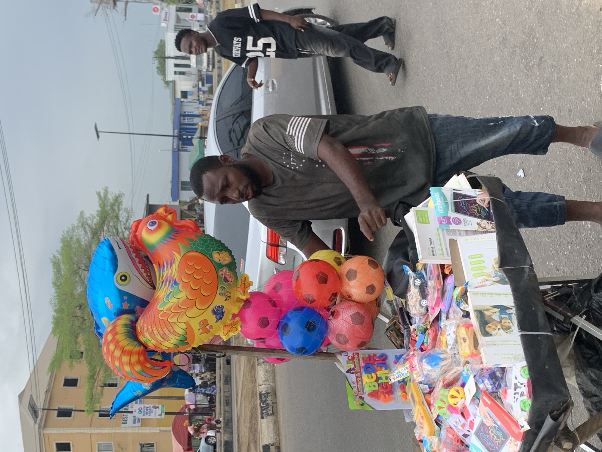 The image shows a street vendor in what appears to be Ikorodu, Nigeria, based on the signage in the background. He stands behind a mobile cart laden with various goods for sale, including balloons shaped like animals (a blue shark and a colorful bird), soccer balls in a net, and various toys and learning materials such as alphabet letters and writing tablets. The vendor is a dark-skinned man wearing a dark-colored t-shirt and blue jeans. Another man, wearing a black t-shirt with the number 25 and the word "CRRYPAYS" on it, stands in the background beside a silver car. The environment includes buildings, an electrical pole with visible wires, and other vehicles. The street is paved, and the weather appears overcast.