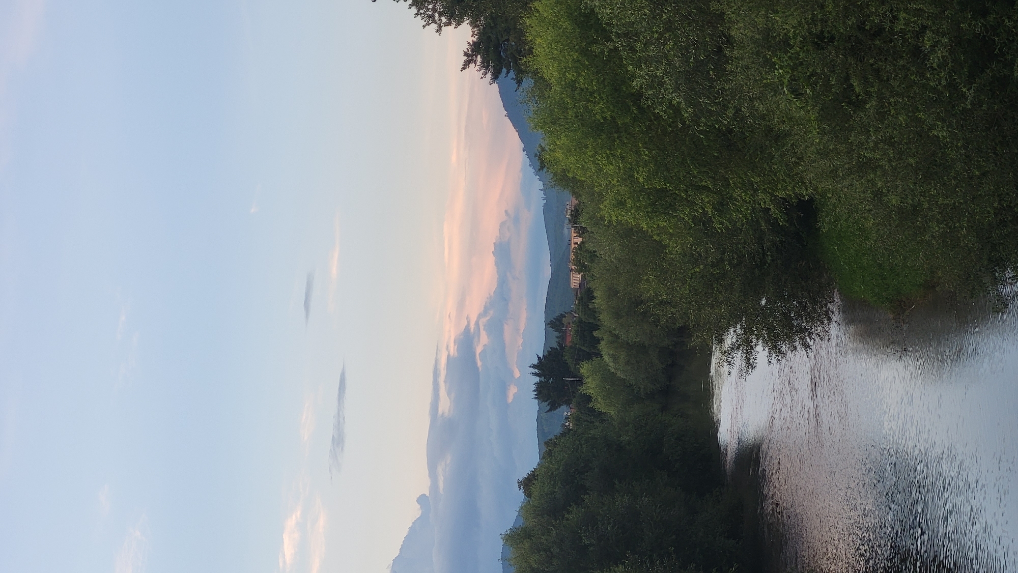 The image depicts a tranquil natural landscape with a river flowing through lush greenery under a soft, pale sky. The river, occupying the lower left portion of the frame, has a calm surface reflecting the muted tones of the sky and surrounding vegetation. The banks of the river are densely lined with various trees and shrubs, their foliage creating a rich, green canopy that extends into the middle ground. In the distance, rolling hills form the horizon, with subtle layers of atmospheric haze softening their contours. Scattered across the sky are wispy clouds, some tinged with the soft hues of either sunrise or sunset, indicating the time of day. The overall impression is one of peaceful, natural beauty. There are no people or specific urban features visible that would definitively place this scene in Prešov, Slovakia.