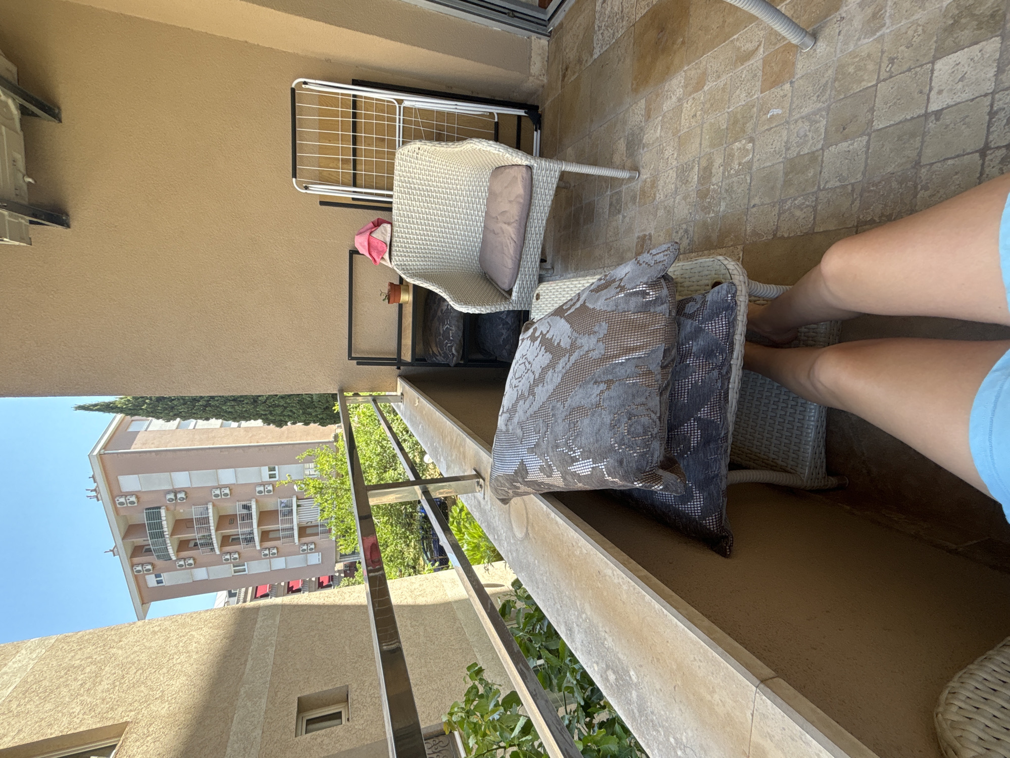 The image depicts a balcony in Budva, Montenegro, with a view of buildings and trees. On the balcony, there is a white wicker chair with a cushion, a small plant on a shelf, and a drying rack. Two decorative pillows are stacked on a small wicker stool in the foreground. A person's legs are visible, resting on the stool, wearing blue shorts. The floor is tiled with square, multi-colored tiles. The balcony has a concrete railing and a textured wall. In the background, a multi-story building with balconies and air conditioning units is visible, along with a tall, slender tree. The sky is clear and blue.