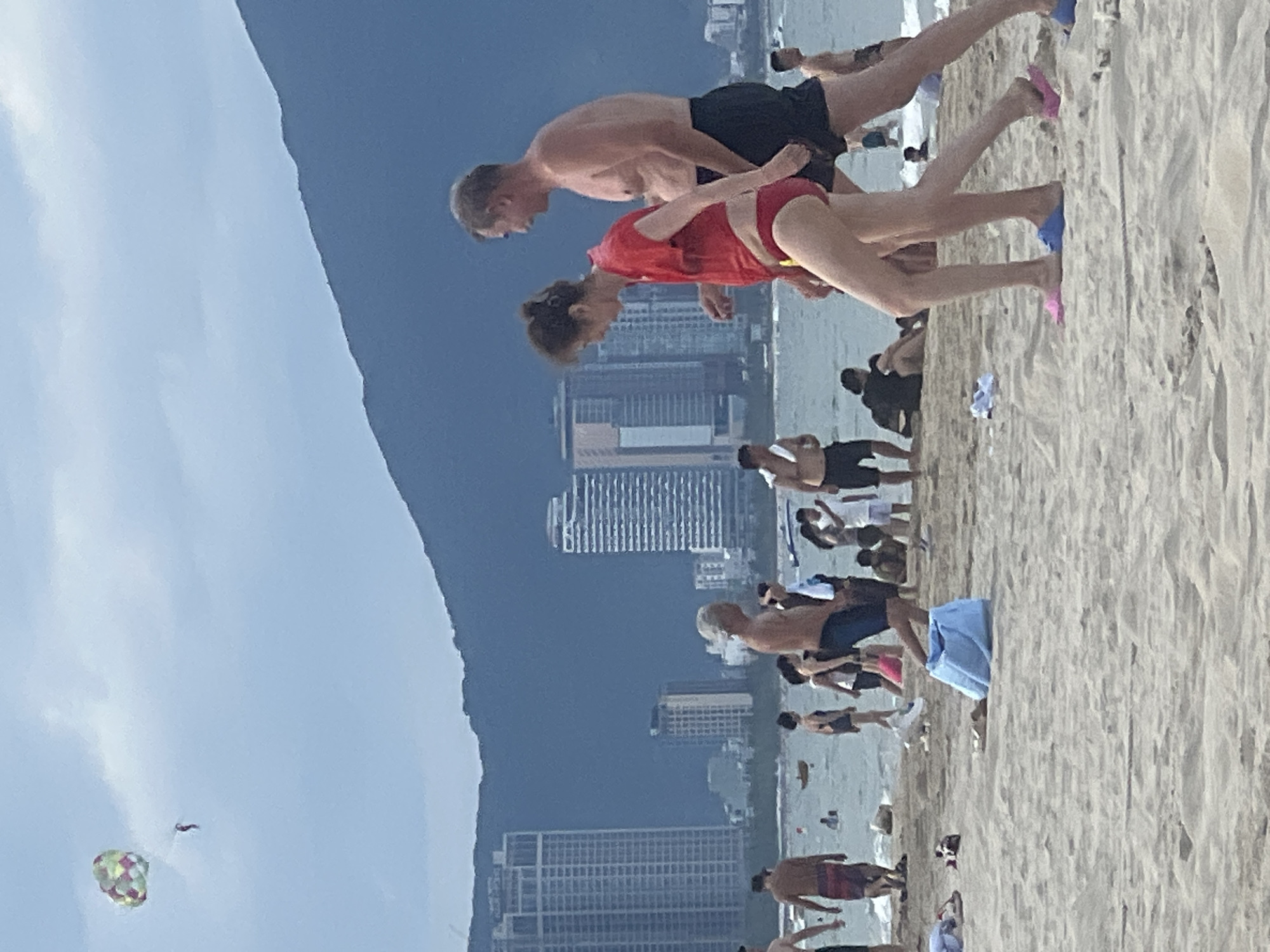 A group of people are enjoying a day at the beach with a city skyline and mountains in the background. The weather is overcast. In the foreground, a couple walks across the sand, the woman in a red swimsuit and the man in black swim trunks. A colorful parachute is visible in the sky, with a person being pulled by it. The beach is dotted with other beachgoers, some swimming in the ocean, others relaxing on the sand. Buildings and palm trees line the shore, and a dark mountain range looms in the distance. The scene is likely located in the city of Đà Nẵng, Vietnam, known for its beautiful beaches and scenic coastal views.
