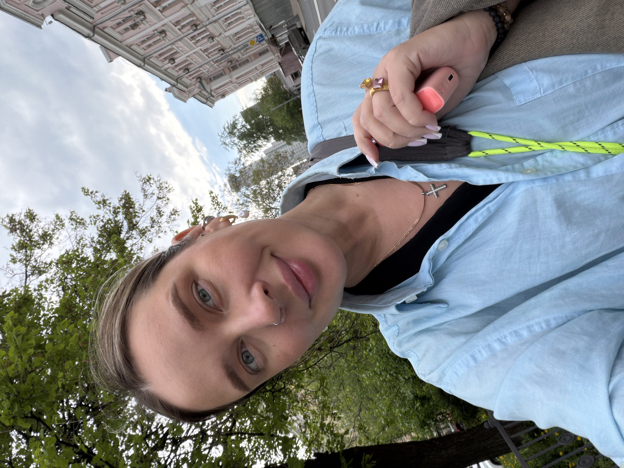 The photograph is a selfie taken by a woman outdoors. She is wearing a light blue shirt, a black top underneath, and a brown jacket. She has a gold necklace with a cross pendant. She is holding a pink device in her right hand and wearing two rings on her right hand. The background includes trees and a building with intricate architectural details, suggestive of Moscow, Russia. The sky is cloudy.