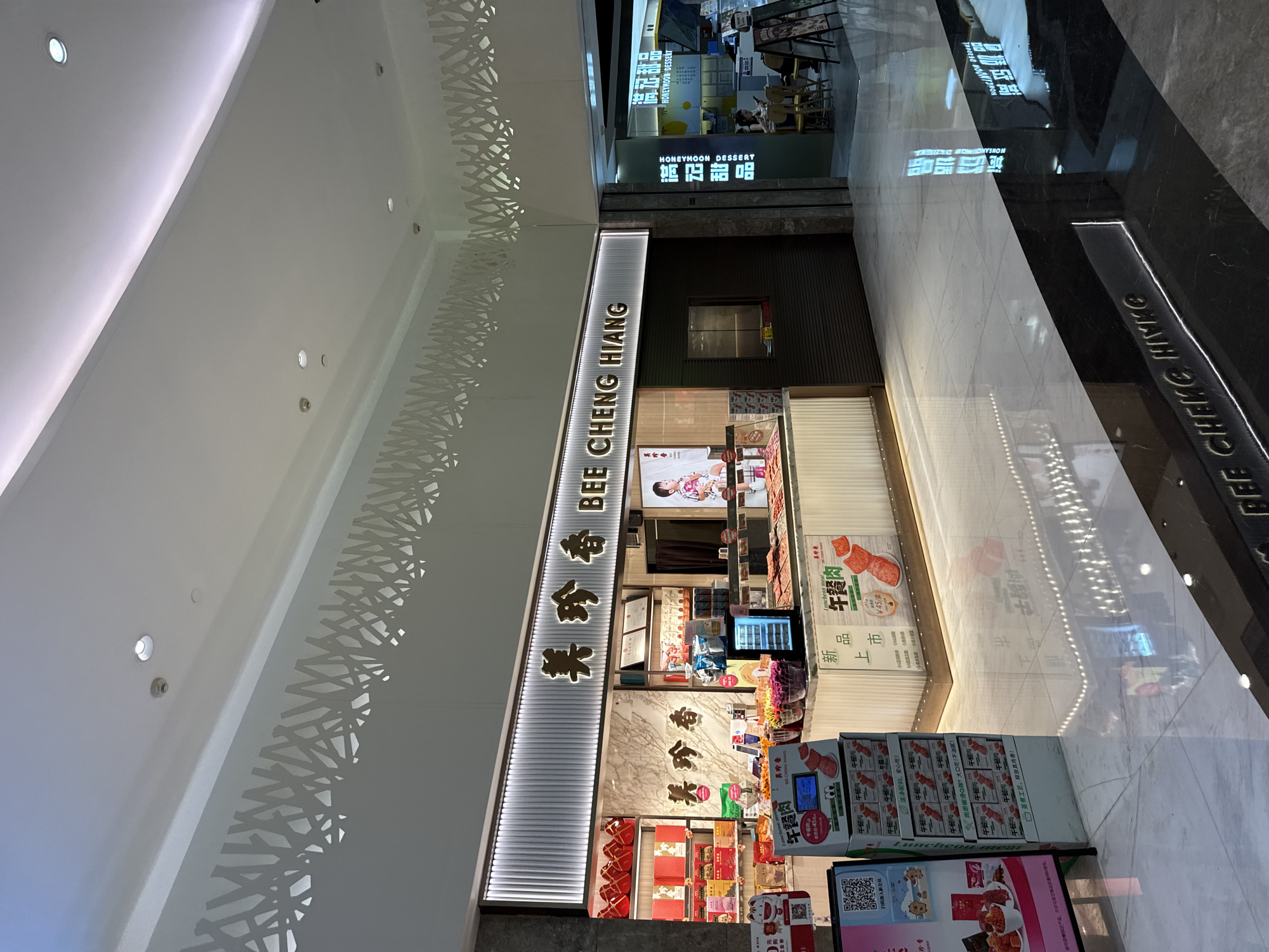 The image captures a retail establishment, identified by signage as "BEE CHENG HIANG," located within a modern, well-lit interior space, likely a shopping mall in Tianhe District, China. The foreground displays a glossy, marbled floor with reflections of overhead lighting and storefronts. To the right, a section of the floor is dark, featuring a glowing illuminated sign that reads "BEE CHENG HIANG" in embossed lettering.
The main subject is the "BEE CHENG HIANG" store, characterized by its clean, contemporary design. The store's facade features a prominent illuminated sign with Chinese characters and the English name. Behind a counter, various products are displayed on shelves and in refrigerated units. These products appear to be packaged food items, possibly dried meats or snacks, with prominent branding and informational displays. There is a large cardboard display unit to the left, showcasing boxes of what appears to be "Lunchmeat" with cartoonish illustrations. Another display, to the right of the counter, features rows of packaged products.
A small, refrigerated display case is visible on the counter, holding samples of the products. Behind the counter, a person is partially visible, seemingly attending to a customer. A screen on the counter displays promotional content. To the immediate right of the "BEE CHENG HIANG" store, another establishment with signage in Chinese and English, "HONEYMOON DESSERT," is visible. This neighboring store has a display of desserts and a seating area where a person is seated at a table.
The overall environment is clean and well-maintained, with a focus on modern retail aesthetics. The lighting is bright and uniform, emanating from recessed ceiling fixtures and the illuminated signage. The presence of multiple retail outlets suggests a commercial or entertainment district.