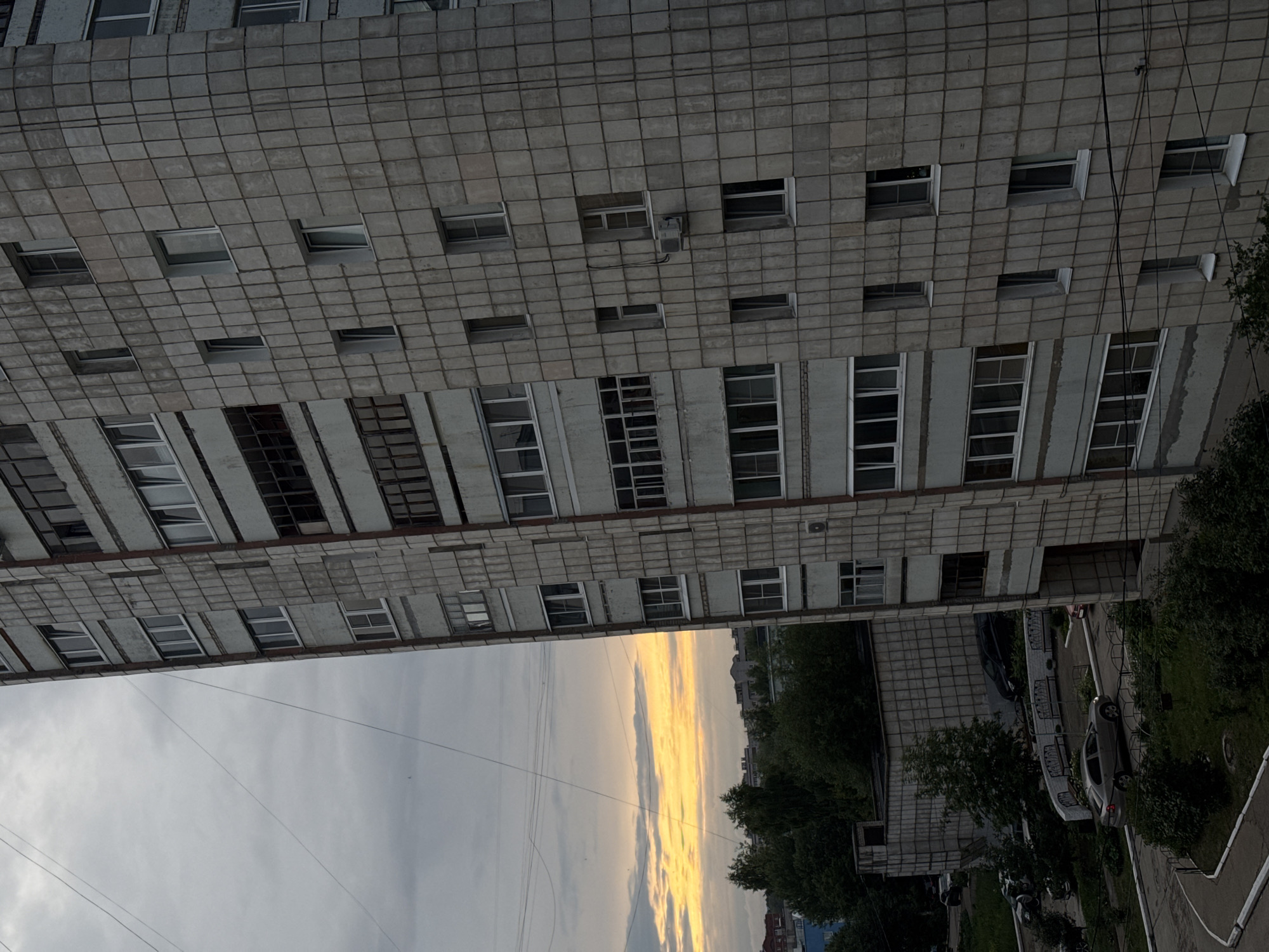 This media file depicts an outdoor scene, likely taken from a high vantage point, of a grey, block-style apartment building in Chaikovsky, Russia. The building's facade is constructed with concrete panels and features numerous windows, many of which are dark or reflect the sky. The image is oriented vertically, making the building appear to extend downwards from the top of the frame.
Below the building, a strip of land is visible, transitioning into a sky with dramatic cloud formations. The clouds are illuminated by what appears to be sunset or sunrise, with streaks of bright yellow and orange light contrasting with the grey and white of the rest of the sky. This suggests the time of day is either early morning or late evening.
The lower portion of the frame shows a street or courtyard area with trees and a few parked cars. The overall impression is one of a somewhat overcast but visually striking sky, juxtaposed with the utilitarian architecture of the apartment block. No people are clearly visible. The scene appears to be a typical urban or suburban residential setting.