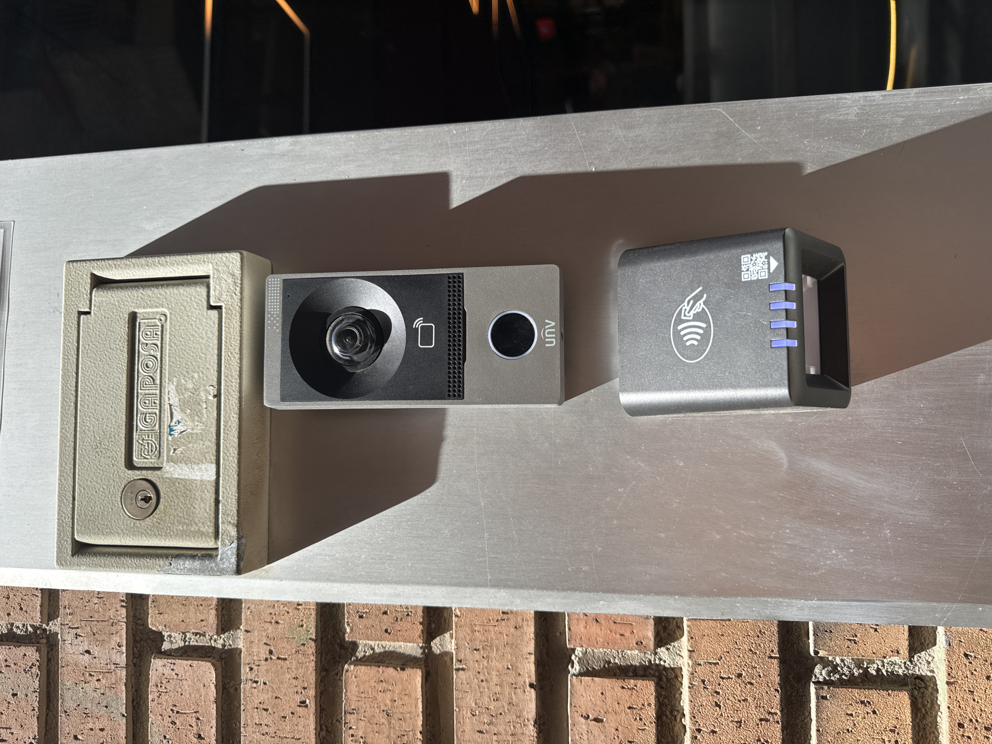 The image shows a close-up of an outdoor security system mounted on a metallic surface, above a brick wall. On the left is a beige, rectangular metal box with a hinged lid, which appears to be a key or intercom access panel. A small circular lock mechanism is visible on the lower portion of this box. To its right is a sleek, silver and black video doorbell or camera unit. It features a prominent lens in the center, with a speaker grille to its right and a circular button below the grille. The word "UNV" is printed vertically on the right side of this unit. Further to the right is a dark gray card reader with a slot at the top. The front of the card reader is adorned with a QR code, a graphic of a hand holding a card with radio waves emanating from it, and a series of three blue LED indicator lights. The metallic surface shows some scratches and dust. The scene is illuminated by artificial light, casting distinct shadows, suggesting it is either nighttime or an indoor area with strong lighting. The brickwork below indicates an outdoor location, likely an entrance to a building. The overall impression is one of modern access control technology. No people are visible in the frame.