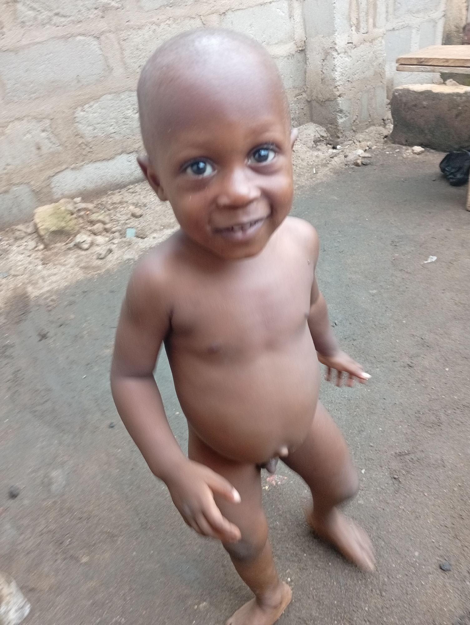 A young, bald, dark-skinned boy stands outdoors on a dirt surface. He is unclothed. In the background is a wall constructed of concrete blocks. A wooden table and debris are also visible. The location is Owerri, Nigeria.