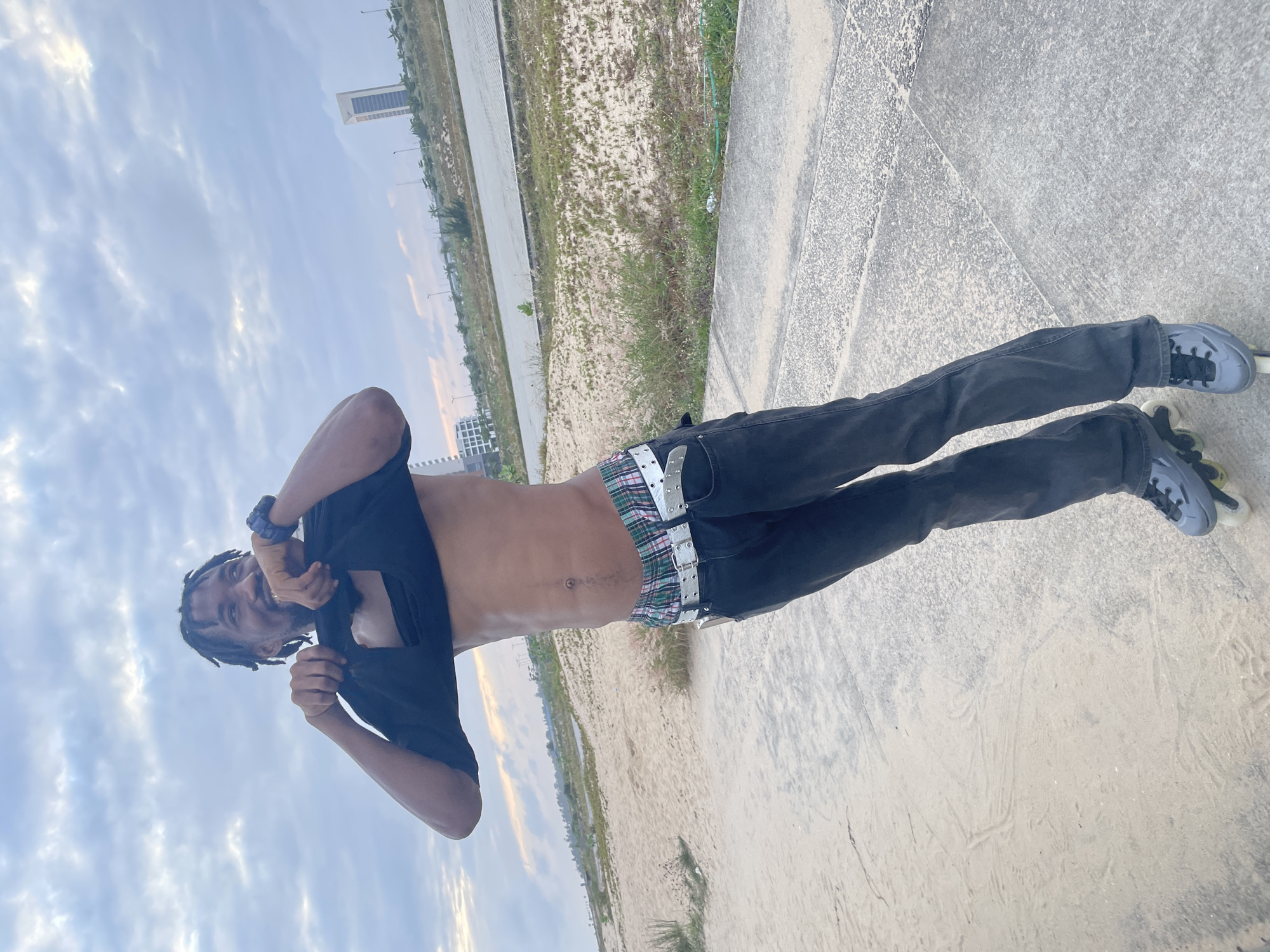 A man with dreadlocks is standing on a paved surface next to a sandy area, pulling up his black t-shirt to expose his bare torso. He is wearing dark-wash flared jeans with a colorful plaid waistband visible, and a silver belt. He has on grey inline skates. In the background, there is a road, some sparse vegetation, and modern buildings, including a tall, white skyscraper in the distance. The sky is cloudy and overcast. The scene appears to be in a coastal area, possibly a promenade or park adjacent to a beach. The location in the city of Inupa Kekere, Nigeria, is suggested by the modern urban development visible in the distance.