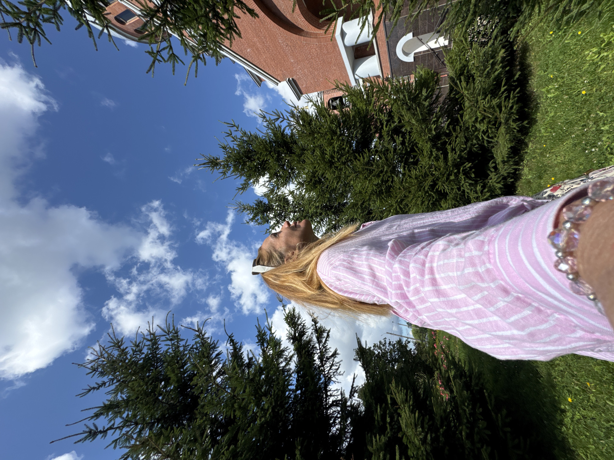 In Sinyavino, Russia, on a sunny day, a woman with blonde hair tied in a ponytail is captured in a profile shot. She is wearing a pink and white striped shirt and a bracelet adorned with pink beads. The woman is looking upwards and to the right. She is positioned outdoors, surrounded by lush green fir trees. In the background, a red brick building, possibly a church, is visible with arched windows and architectural details. The sky is a vibrant blue, dotted with white, fluffy clouds. The ground is covered in green grass with scattered yellow wildflowers.