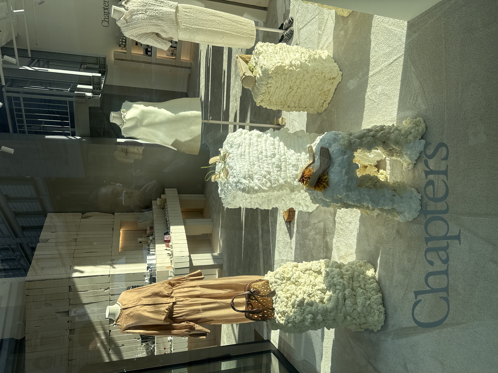 The image shows a storefront display for a brand named "Chapters." Mannequins are dressed in clothing, including a tan dress, a white top, and a white knitted dress. The displays are set against a backdrop of stacked white blocks and textured white surfaces. Some displays include unique textures that are used as abstract decoration and to hold accessories. The storefront is located in a city, most likely Milan, as the store is located in Milan, Italy. The image is taken through a glass window. Reflections on the glass are visible.