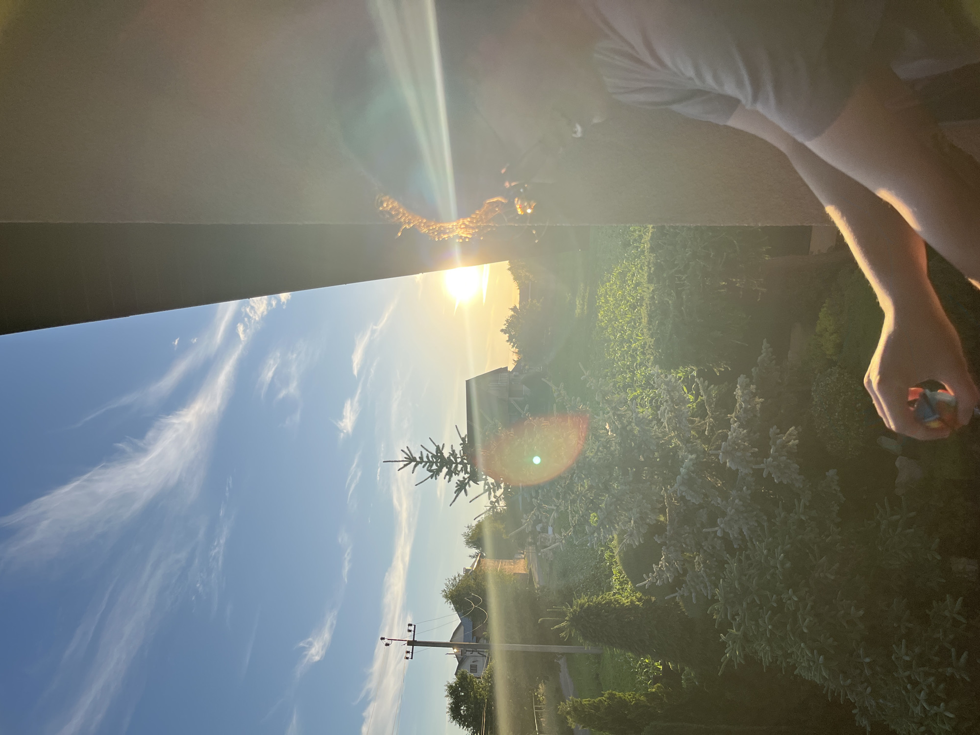 In Koroviia, Ukraine, a person's right arm and shoulder are visible in the foreground, leaning against a light-colored wall. The person is wearing a grey t-shirt and appears to be holding a small, colorful object in their right hand. The background depicts a vibrant sunset over a rural landscape. Tall, wispy clouds streak across a clear blue sky. The setting sun casts a warm, golden glow over the scene, creating lens flare effects. Green trees, including a tall evergreen, and a small building with a dark roof are visible. A utility pole with wires stands to the left of the frame. The overall environment suggests an outdoor setting during the golden hour.