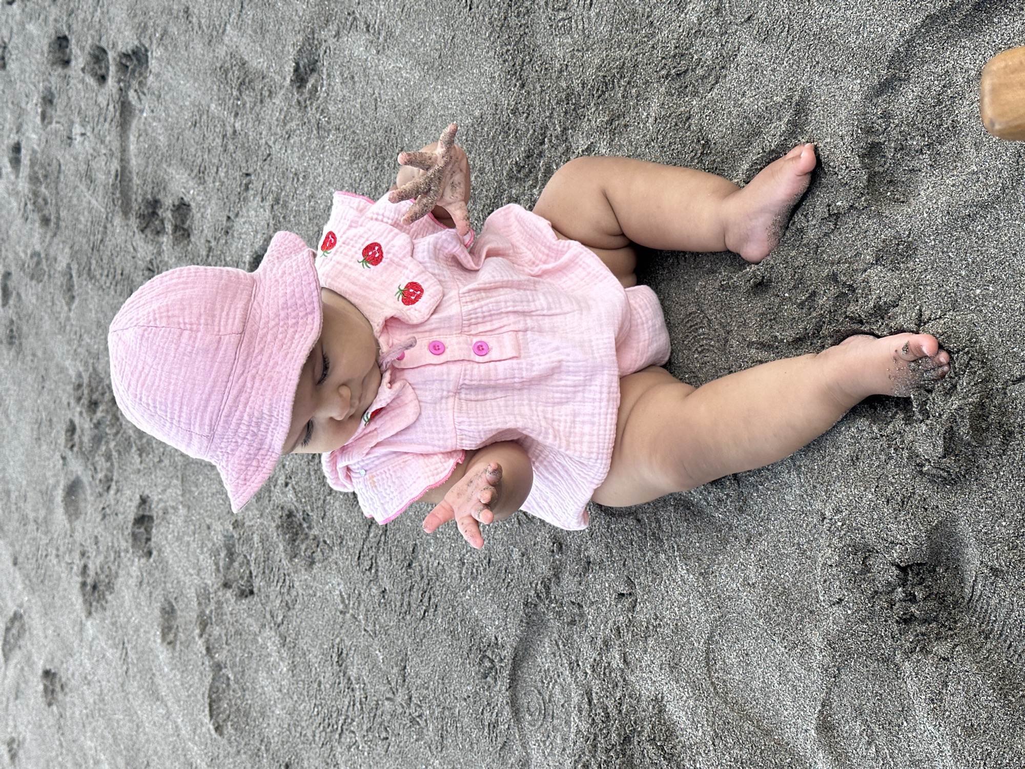 A baby lies on a sandy beach. The infant wears a pink sun hat and a matching pink strawberry-themed shirt. Sand clings to the baby’s hands and feet. The beach is dark gray. The location is North Kuta, Indonesia.