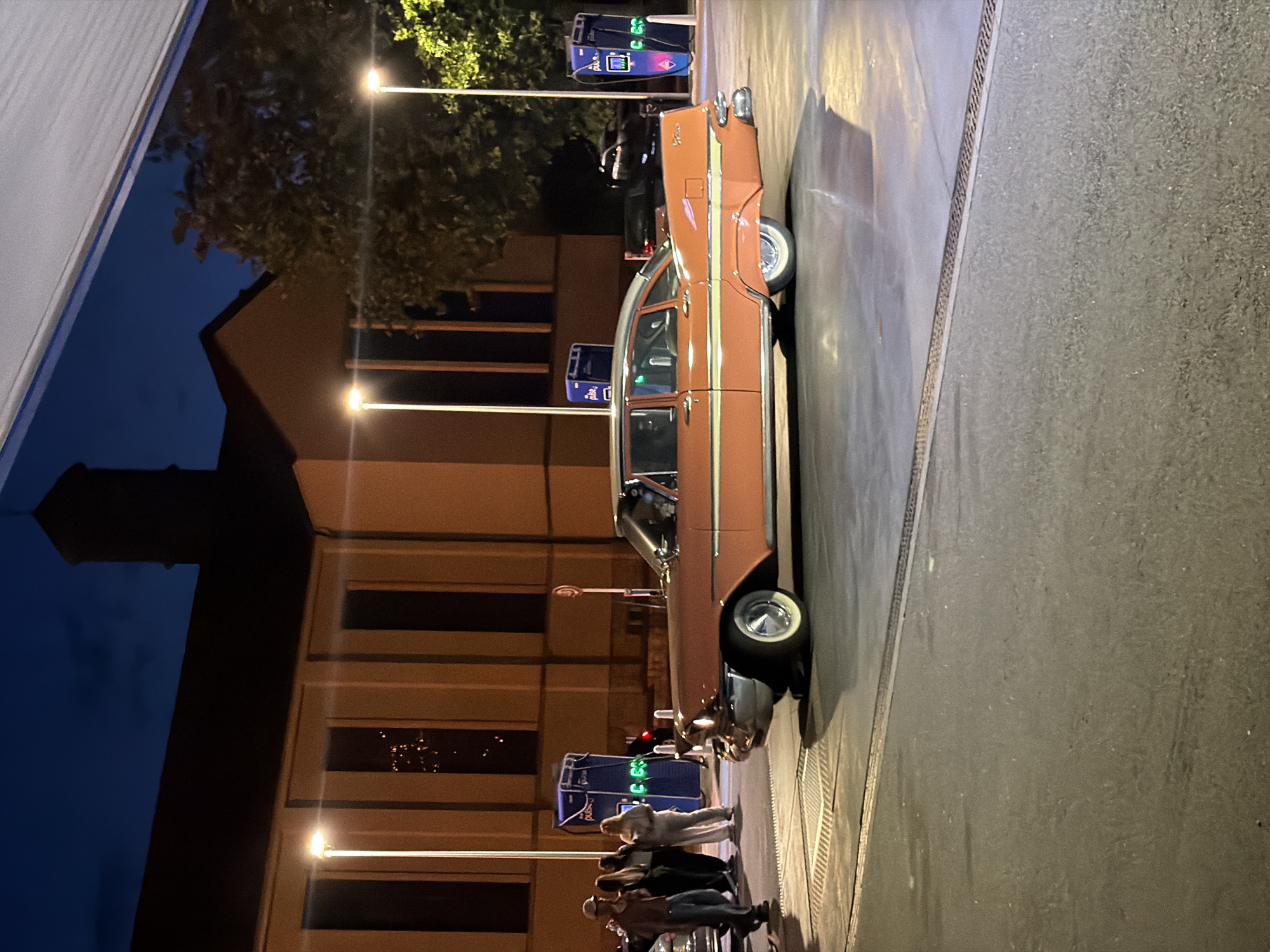 At dusk, under a deep blue sky, a vintage salmon-pink car is parked on a wet asphalt surface. The car, a classic sedan with chrome accents and whitewall tires, is positioned to the right of the frame. Behind the car, a building with a dark, sloped roof and tall, narrow windows is visible, illuminated by streetlights. Several people, dressed casually, stand near some blue charging stations, suggesting a public charging point or a service station. The scene appears to be in an urban environment, possibly Esch-sur-Alzette, Luxembourg, given the presence of what appear to be charging stations that may be part of a modern infrastructure. The lighting suggests artificial illumination from streetlights and the charging units, creating reflections on the wet ground.