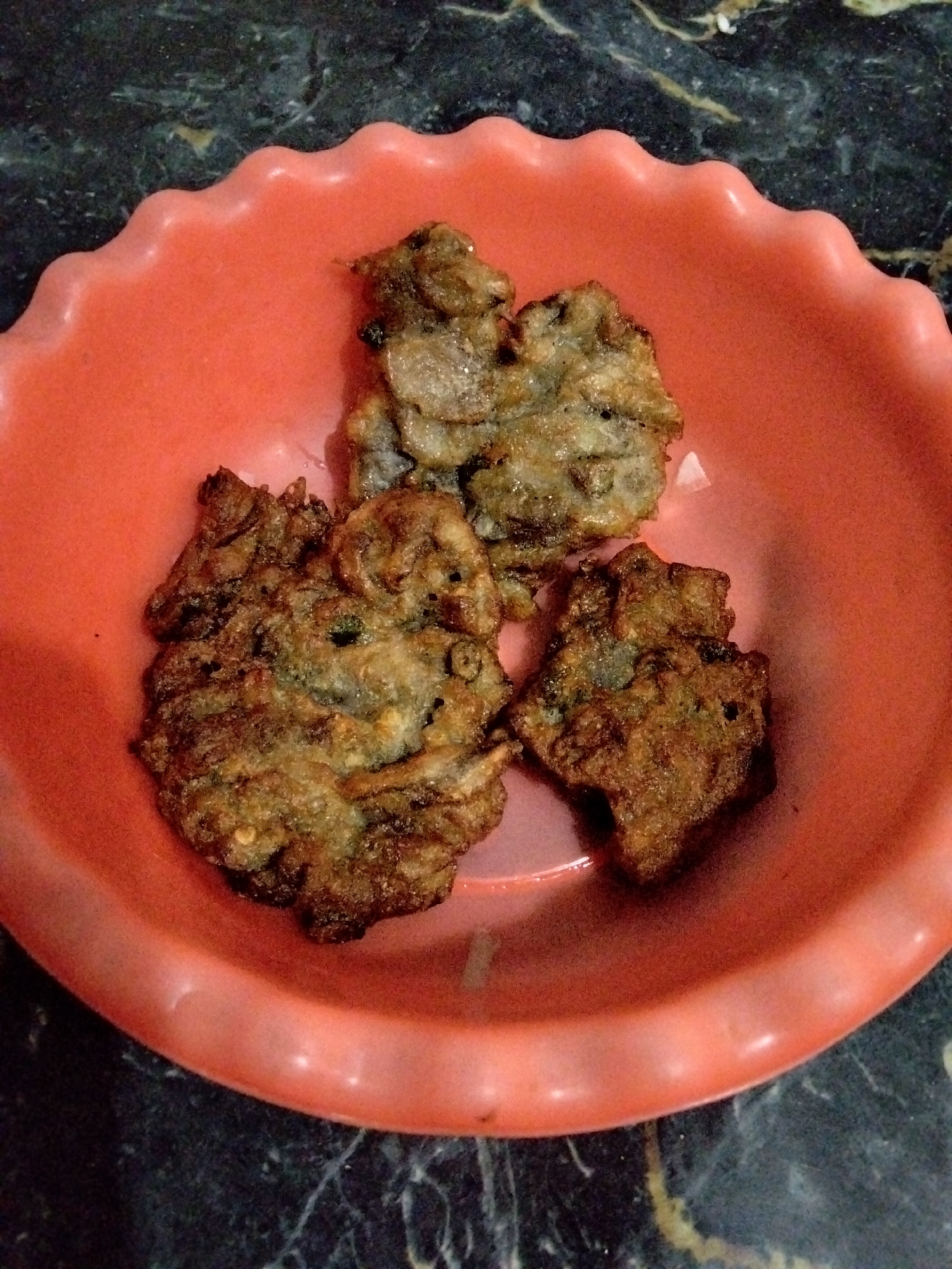 A close-up shot captures three savory fritters, appearing to be of Indonesian origin, served in a fluted coral-colored bowl. The fritters are irregularly shaped and golden brown, suggesting they have been fried. They are nestled in the center of the bowl, which is placed on a dark, marbled surface. The lighting is somewhat dim, casting a slight shadow. The background is out of focus, but shows the continuation of the marbled surface. There is no text visible in the image. The scene is likely indoors, possibly in a home kitchen or dining area. The fritters themselves appear to be a popular street food or snack, common in Indonesian cuisine. The image has a warm, inviting feel, highlighting the appetizing appearance of the food.