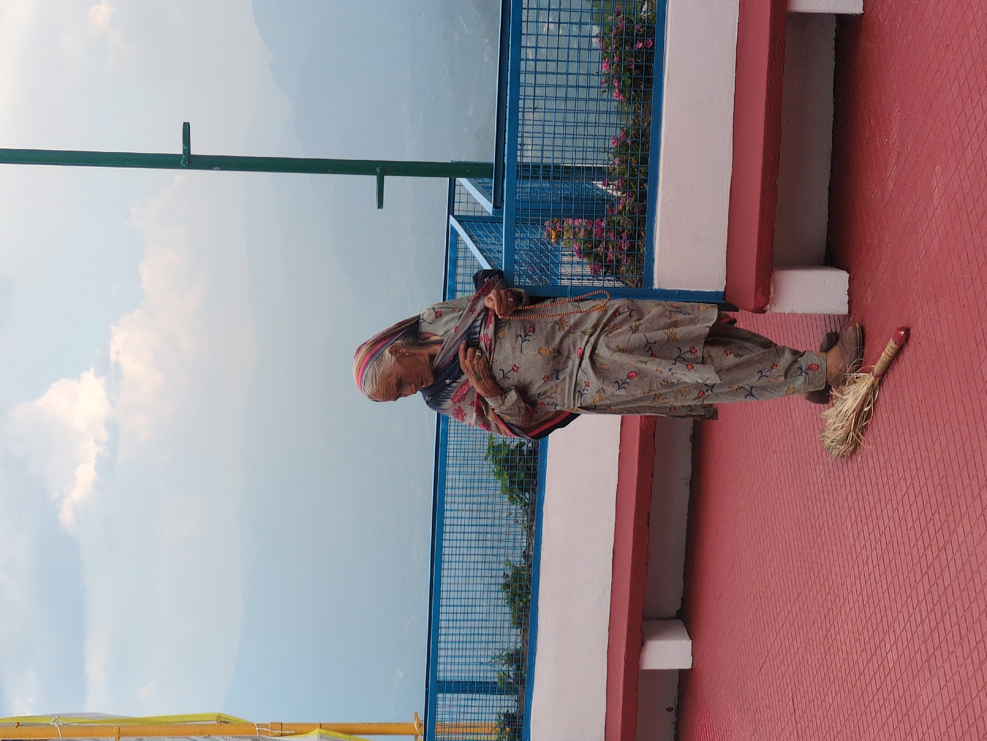 An elderly woman stands on a red, textured outdoor platform, holding rosary beads and wearing a traditional gray salwar kameez with a patterned shawl draped over her shoulders. A broom lies on the ground in front of her. She is positioned near a white and red bench and a blue metal fence that encloses a garden with pink flowers. The background shows a hazy mountainous landscape under a cloudy sky, suggesting a high-altitude location. The setting appears to be a public space or a viewing area. The overall atmosphere is serene and contemplative. This scene is not located in Doh, India, as there is no city named Doh in India.