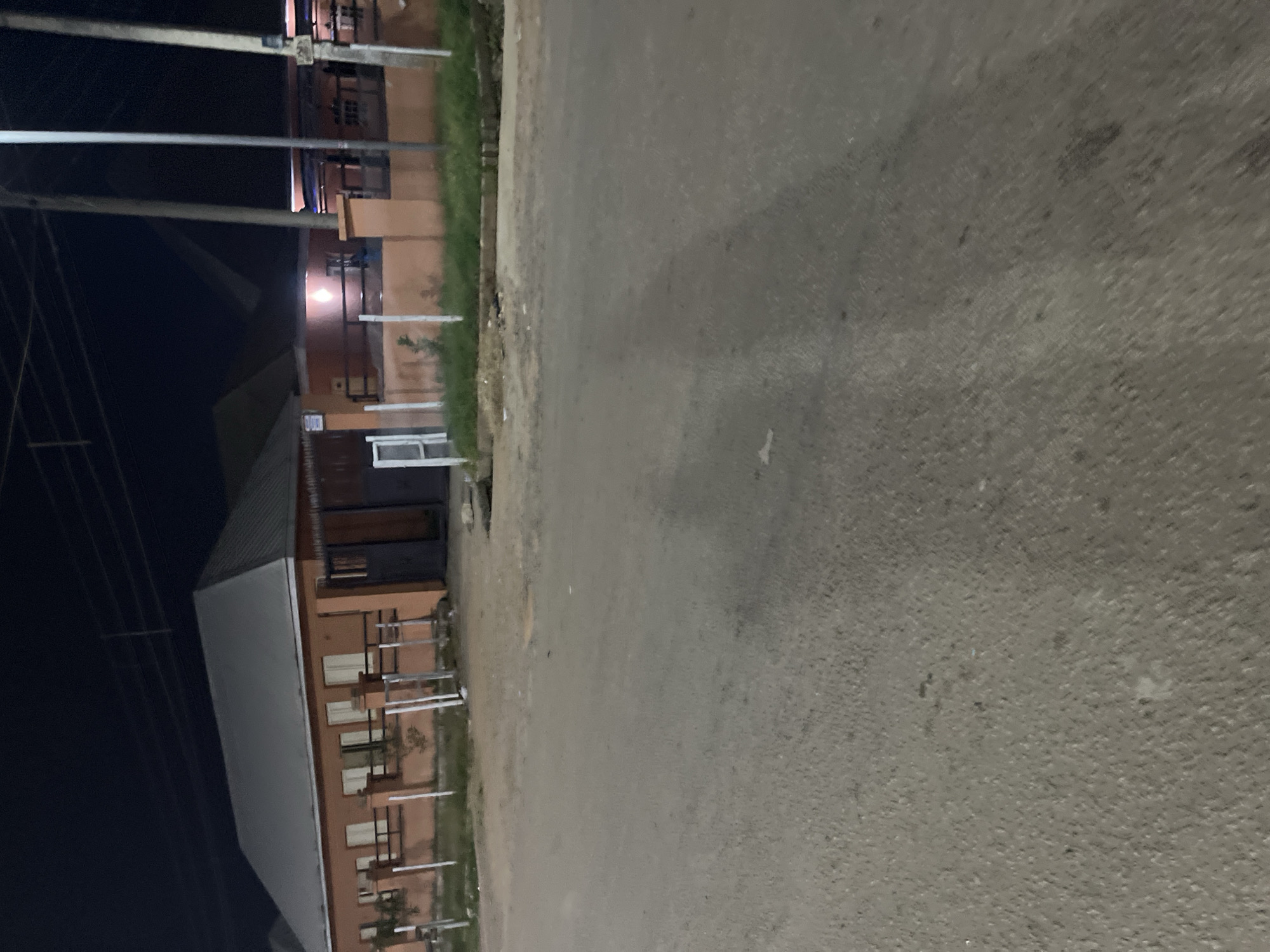 The image depicts a street scene at night in Abuja, Nigeria. In the background, a single-story building with an orange facade and a gray pitched roof is visible, bordered by a fence. The building has several windows with cream-colored coverings. A gate is present on the right side of the building. Several utility poles stand in front of the building, and power lines are strung across the dark sky. The foreground is dominated by a paved road, showing signs of wear and tear, with some scattered debris and a manhole cover. The lighting suggests artificial illumination from streetlights or the building itself.