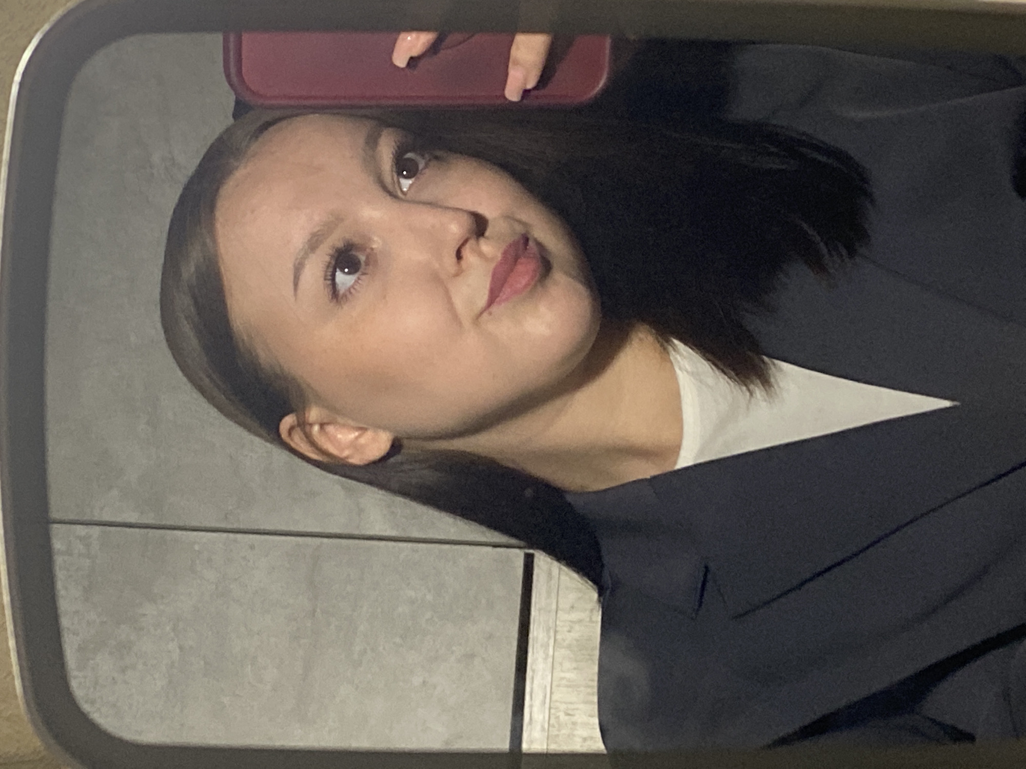 An indoor, mirror selfie captures a young woman from Chaikovsky, Russia, in a close-up, chest-up shot. The image is rotated counter-clockwise, showing her head tilted upwards and to the viewer's right, her gaze directed similarly. She has dark hair, likely styled straight, and prominent dark eyes. Her lips are adorned with a rosy or muted red lipstick, and she has a thoughtful or slightly pensive expression, with a subtle upward curve to her mouth.
She is wearing a white top, possibly a t-shirt, layered under a dark, possibly navy or black, blazer or jacket. The upper part of a hand, holding a smartphone with a red case, is visible in the reflection near her forehead, indicating she is taking the photo. The reflection also shows a light grey, textured wall, possibly tiled, behind her. The overall lighting appears artificial, casting some shadows and highlights on her face and hair. The scene is framed by the dark, rounded edges of a mirror. There is no visible text in the image.