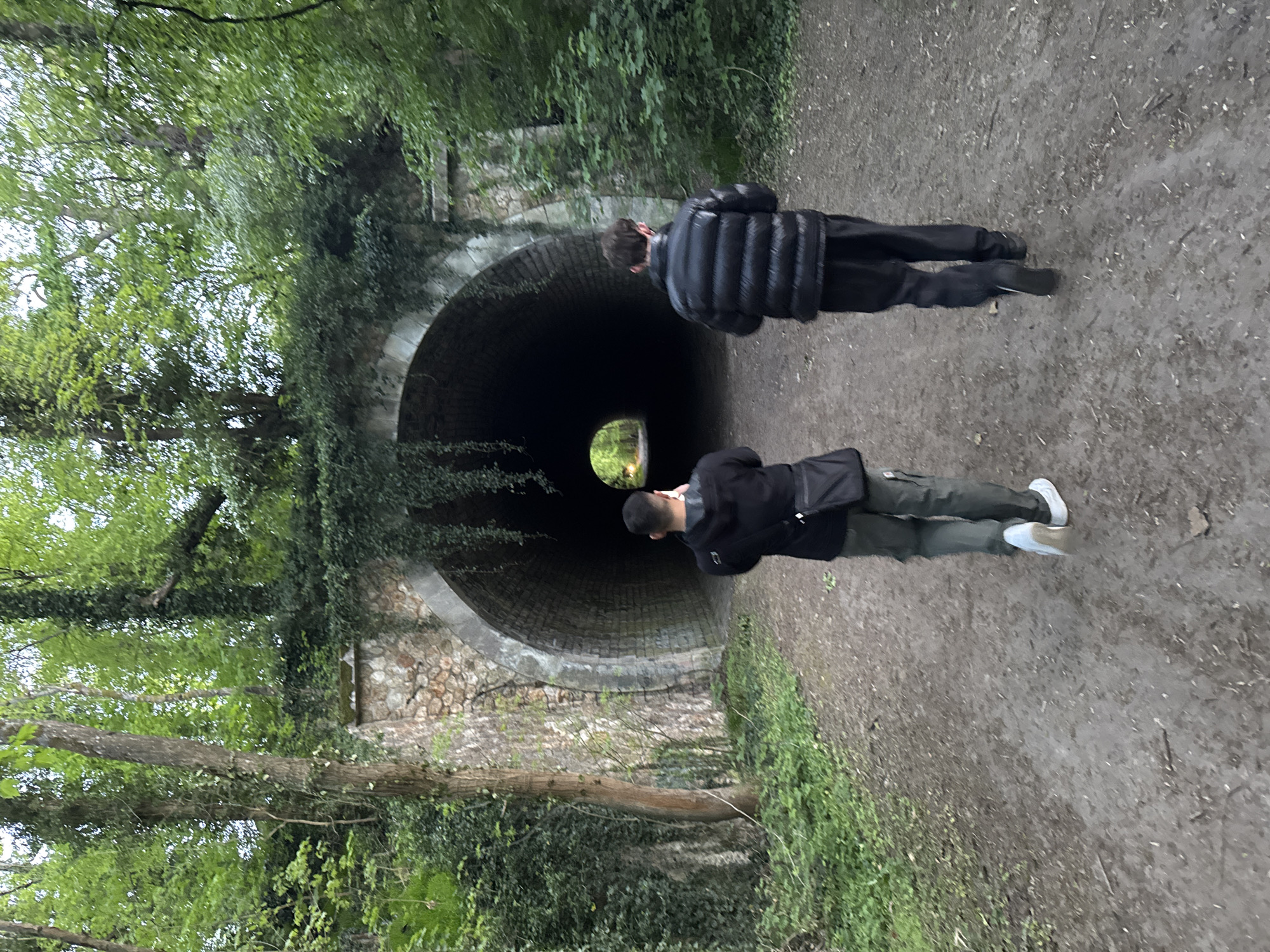 The image depicts an outdoor scene in Bures-sur-Yvette, France, showing two individuals on a dirt path approaching a stone-arched tunnel entrance. The primary subject is the tunnel, which appears dark inside, with a small, bright, circular opening visible at its far end, revealing green foliage and suggesting an exit.
One person is positioned closer to the camera, seen from behind, wearing a dark jacket (possibly black), dark trousers (resembling cargo pants), and white sneakers. They have short, dark hair and are looking towards the tunnel. A dark shoulder bag is slung over their shoulder. The second individual is further away, also facing the tunnel, dressed in a black puffer jacket, black trousers, and dark shoes.
The ground is a well-trodden dirt path, showing some dampness and small stones. The tunnel entrance is constructed from light-colored stone, partially covered in ivy and other green foliage. Dense trees and lush green undergrowth frame the path and the tunnel, creating a natural, somewhat wild setting. The lighting is soft and diffused, indicating an overcast day or shaded area, with brighter light visible at the tunnel's exit. The scene conveys a sense of exploration or passage through a natural landscape, likely a park or forest trail. There is no visible text in the image.