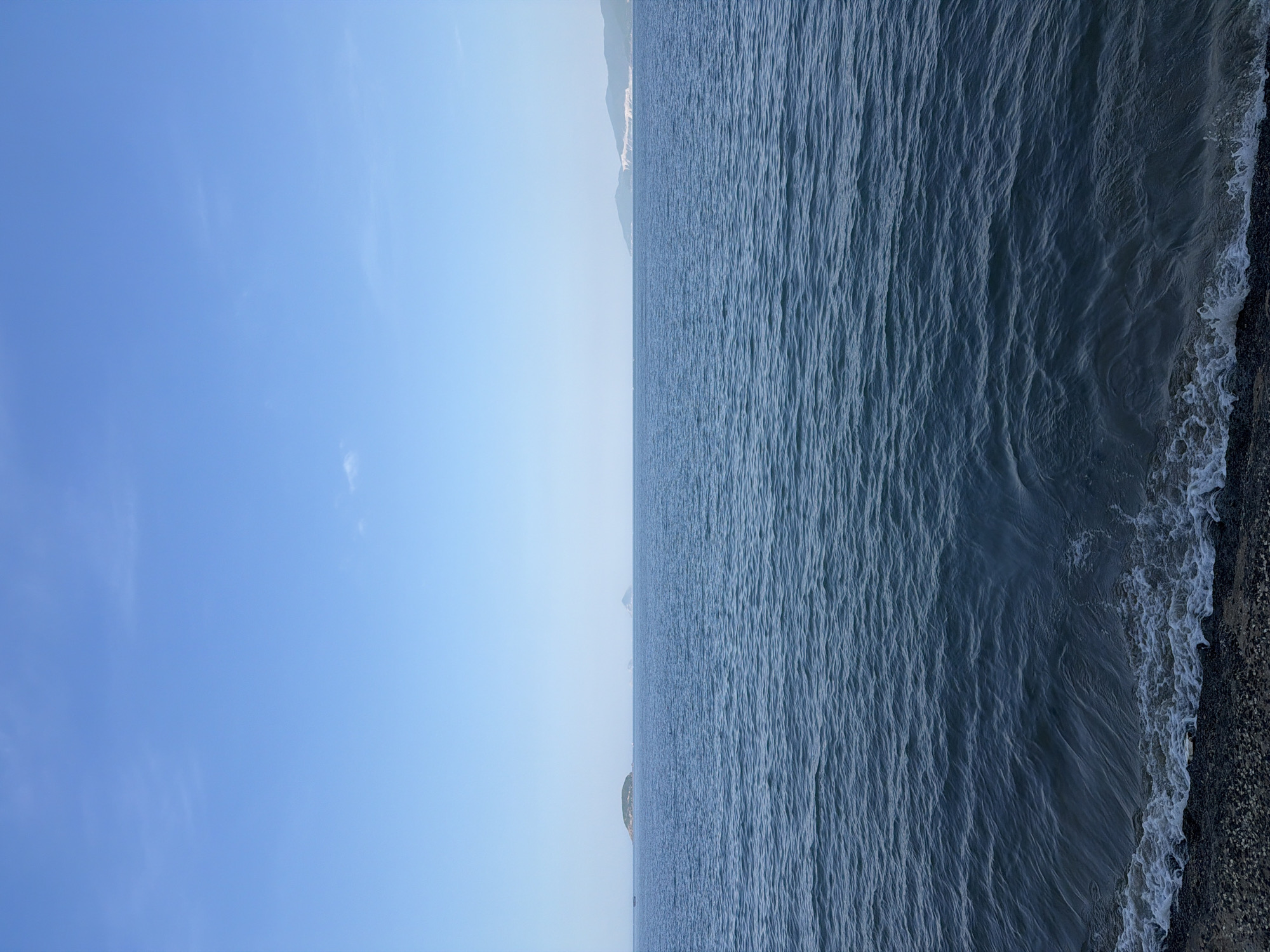The image displays a serene seascape with a clear blue sky dominating the upper portion. The horizon is visible, separating the sky from the ocean. Several small islands are scattered across the water, with one larger, green island in the mid-left of the frame. In the distance, on the right side, a coastline with hills and some structures is faintly discernible. The ocean is calm, with gentle ripples on its surface. The foreground shows a rocky shore with small waves lapping at the edge. The scene appears to be captured from a shoreline in Bac Nha Trang Ward, Vietnam, on a clear day. There are no people or animals visible in the image.