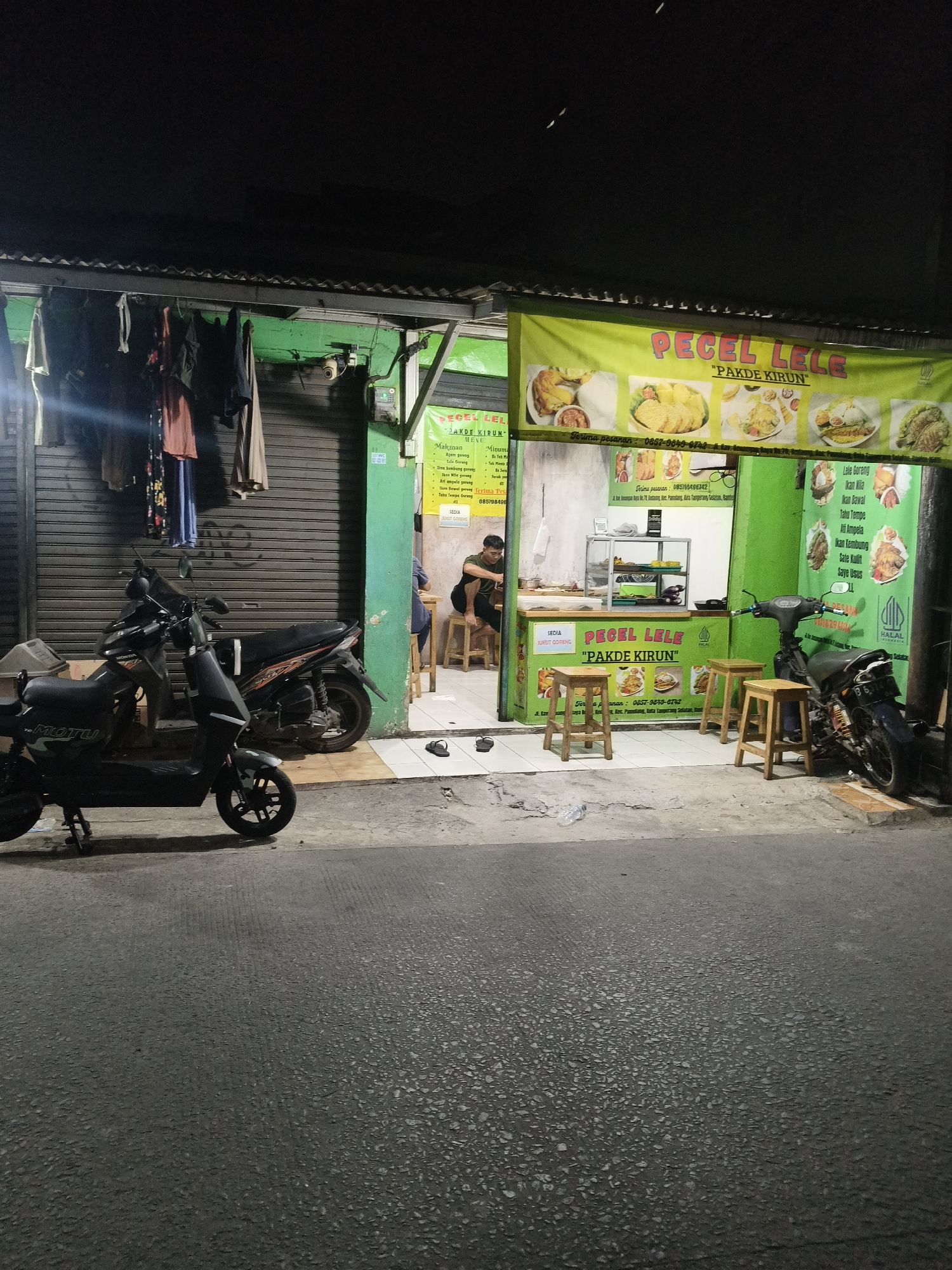 The image depicts a street-level view of a "Pecel Lele" restaurant named "Pakde Kirun" located in South Tangerang, Indonesia. The restaurant features a green facade with a yellow banner displaying images of food and the restaurant's name. Inside, there's a counter with food displays and a person sitting on a stool. In front of the restaurant, two motorcycles are parked, and several wooden stools are arranged for outdoor seating. The street in the foreground is paved with asphalt. The scene appears to be at night, as indicated by the dark sky and the lighting from the restaurant.