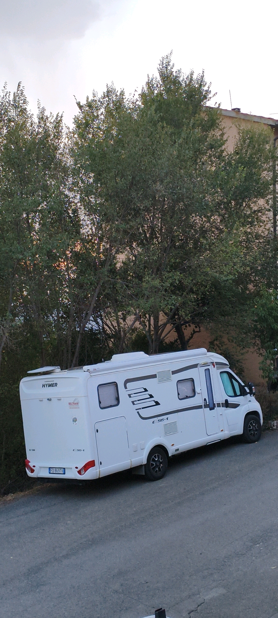 An exterior, low-angle, slightly right-tilted, eye-level shot captures a white recreational vehicle (RV) parked on an inclined asphalt road in Campagnatico, Italy. The RV, a Hymer model, is positioned diagonally from the lower left to the upper right of the frame, facing right. It features multiple windows, an awning, and prominent branding, including "HYMER" and "BELTRANI" on the rear. A license plate reading "FD 869TA" is visible on the back.
Behind the RV, dense green trees with visible branches dominate the left and central portions of the image, obscuring a building that appears to be to the right of the trees. The building's facade is a light brown or peach color, with a dark tiled roof visible above the trees. The sky is overcast with a light gray hue. The ground is paved with asphalt and shows some cracks and wear. There are no people or animals visible in the scene. The overall impression is a still, static shot of a parked RV in a semi-urban or rural setting.