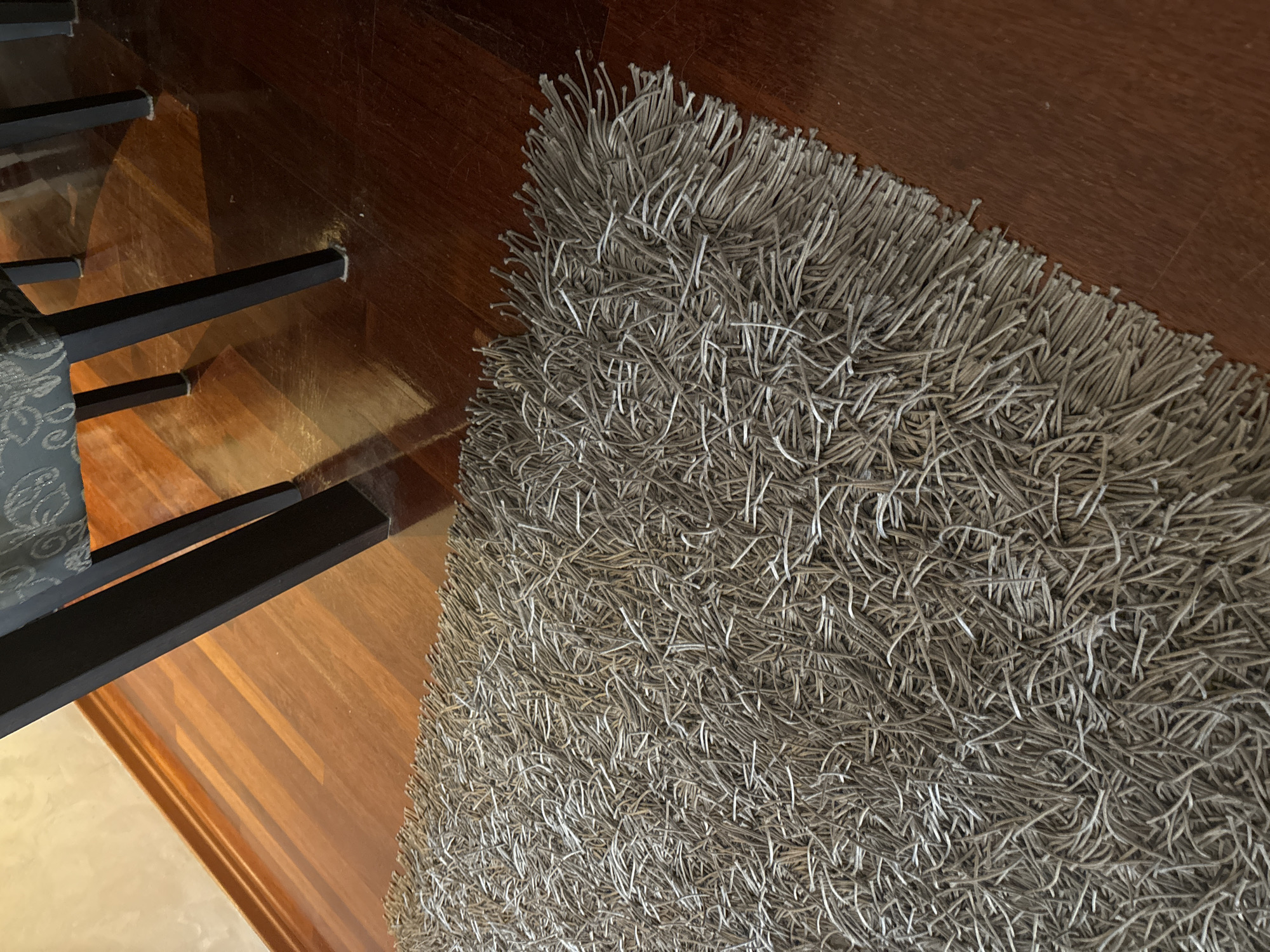 The image shows a close-up view of a shaggy, grey rug placed on a polished wooden floor. The rug's texture is characterized by long, thin strands that create a dense, fuzzy appearance. The wooden floor has a warm, reddish-brown hue and reflects light, indicating a glossy finish. In the background, the dark legs of a chair or table are visible, casting shadows on the floor. The lighting suggests an indoor environment, possibly a living room or dining area. The image is an eye-level shot, focusing on the texture of the rug and the sheen of the floor. No people or specific landmarks are visible.