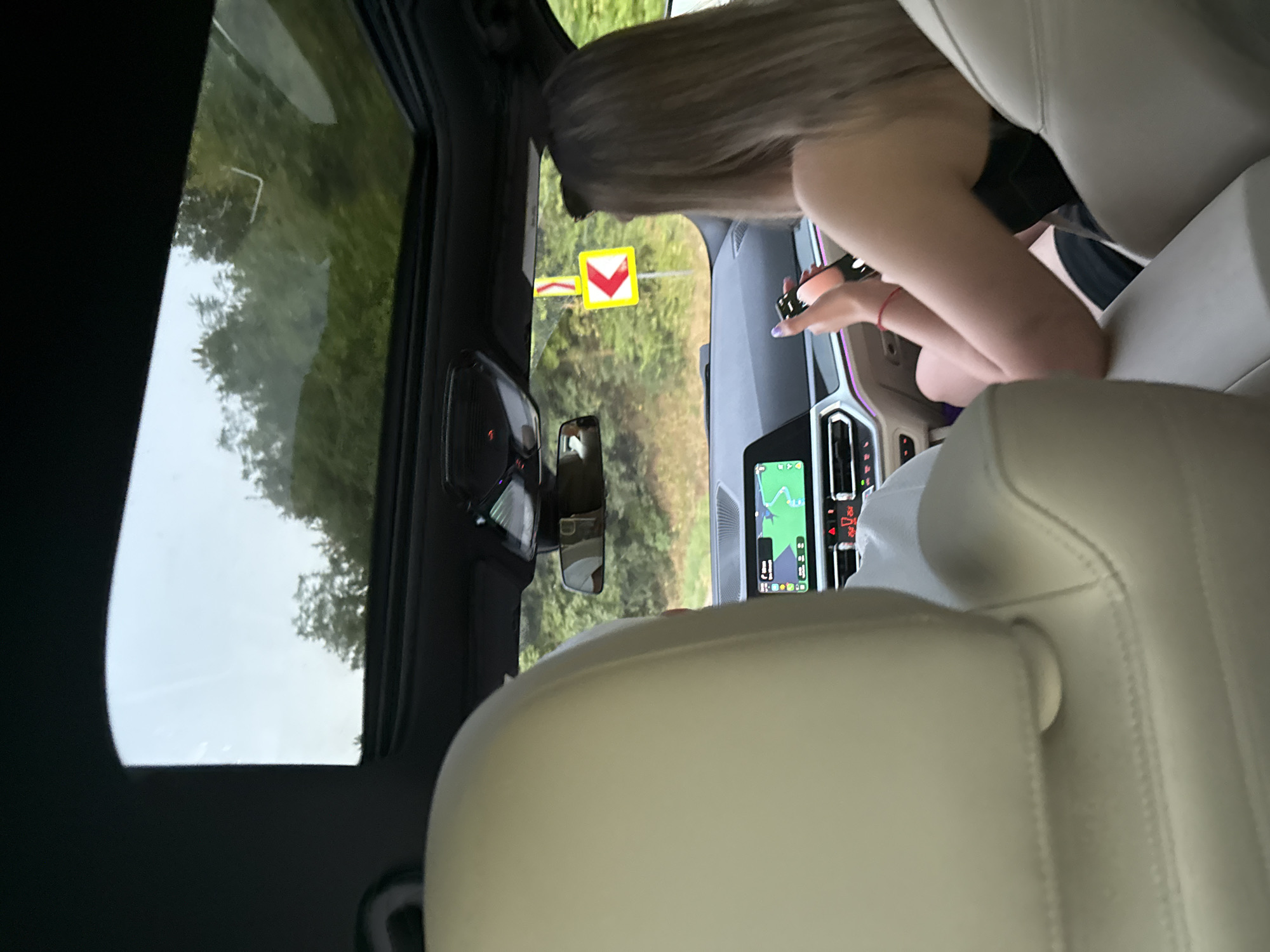The image shows the interior of a car looking forward through the windshield and sunroof. A person with blonde hair is in the driver's seat, looking at their phone. They are wearing a black top and have purple nail polish. The car's navigation system is visible on the dashboard. Outside, a yellow traffic sign with a red border indicates a sharp left turn. Trees and a cloudy sky are visible through the sunroof. The car appears to be on a road surrounded by trees, suggesting a rural or scenic location. The car's interior is upscale, with beige leather seats. The image was taken in Fundul Galbenei, Moldova.