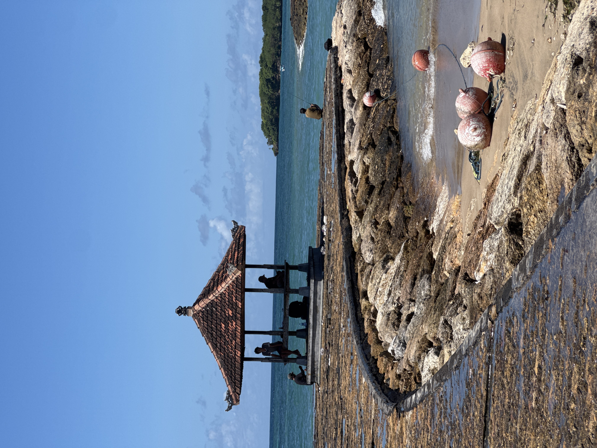The image shows a seaside scene in Kuta Selatan, Indonesia. A traditional Balinese pavilion with a tiered red-tiled roof stands on a concrete platform at the water's edge. Several people are visible within the pavilion. Adjacent to the platform is a rocky breakwater line with orange buoys attached. Beyond the breakwater is a small sandy beach. In the distance, a forested island is seen on the horizon, with the clear blue sky above. A person is fishing off of the breakwater.