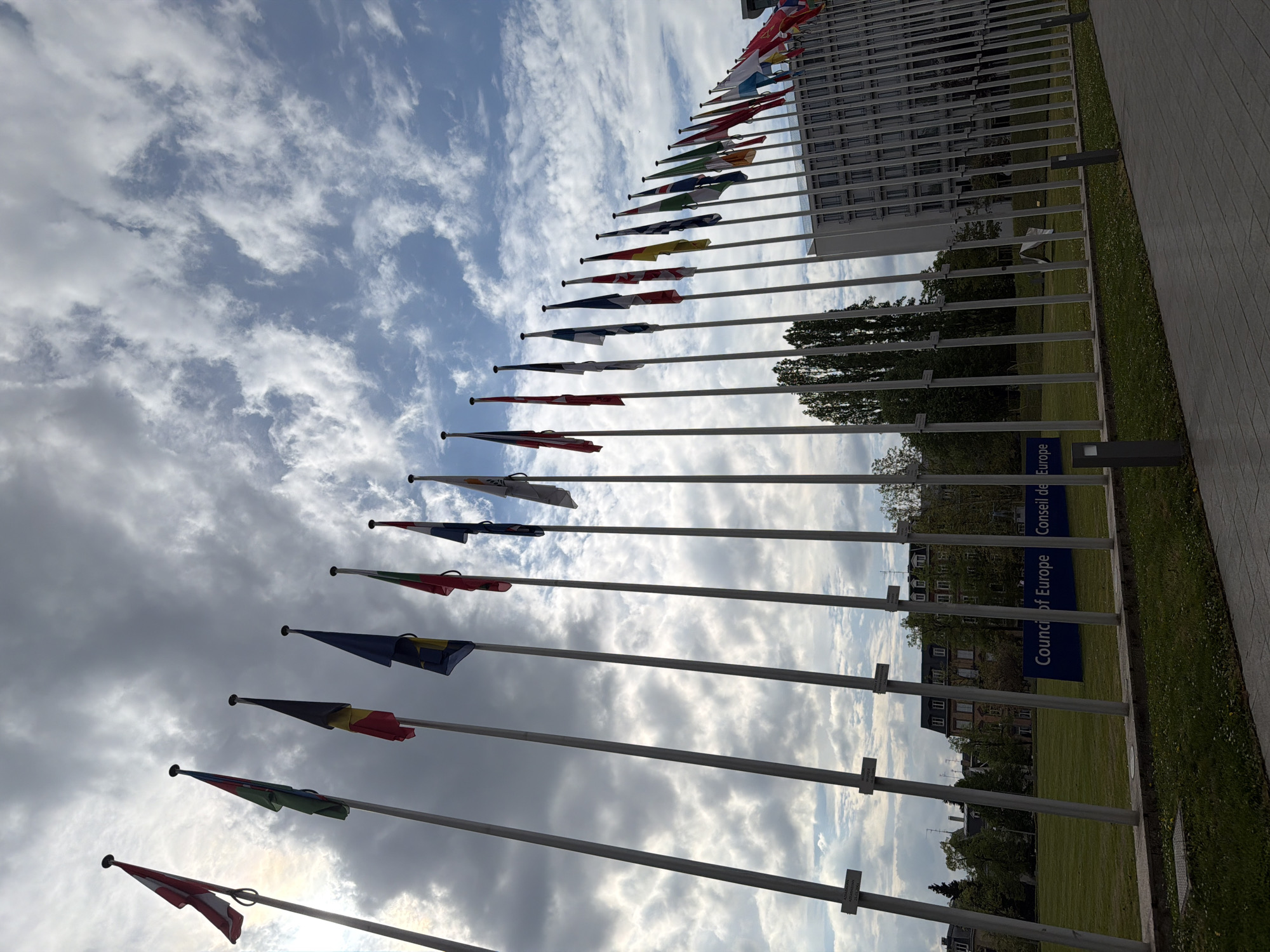 The image captures a striking view of numerous national flags displayed on tall flagpoles, set against a dramatic cloudy sky. The flags, in various colors such as red, white, blue, yellow, green, and black, are partially unfurled, suggesting a light breeze. They extend in a long, receding line from the lower left to the upper right of the frame, leading towards a large, modern building in the background.
The sky is a prominent feature, filled with white and grey cumulus clouds, with occasional patches of bright blue visible through the breaks in the cloud cover. The lighting indicates it is daytime.
In the upper right background, a multi-story, modern institutional building with a repetitive window pattern and a light-colored facade is visible. Below the building and flags, a well-maintained green lawn stretches out, dotted with several deciduous trees. A paved pathway runs along the right edge of the frame.
A key detail identifying the location is a dark blue, vertically oriented sign positioned near the pathway and lawn. It clearly displays the text "Council of Europe" and "Conseil de l'Europe," indicating the setting as the Council of Europe in Strasbourg, France. The overall scene presents a formal and institutional atmosphere under a dynamic sky. There are no people visible, and no specific event or activity is taking place beyond the static display of flags.