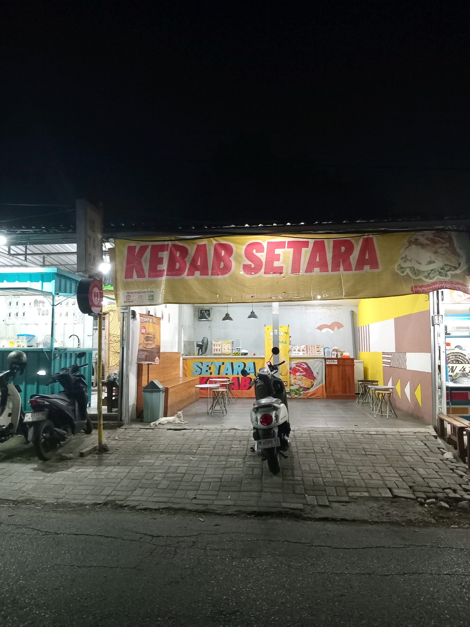 In Randuagung, Indonesia, a night scene captures the storefront of "KEBAB SETARA". A large yellow banner with red lettering announces the establishment. Two scooters are parked on the left side of the paved walkway in front of the shop. A white cat is resting on the pavement in front of the shop. The shop itself is open, with tables and chairs set up inside and outside for customers. A cashier counter is visible inside, and promotional posters with food images adorn the walls. The overall atmosphere suggests a casual, late-night dining spot.
