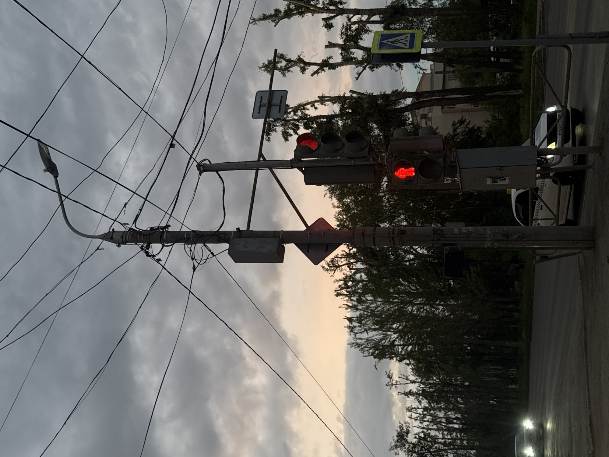 The image shows a street scene in Perm, Russia. The view is tilted counter-clockwise. A traffic light displays a red light and a red arrow. Power lines crisscross the sky. Street signs are visible. Cars are on the road. Trees line the street. The sky is overcast. A street lamp is attached to a utility pole.