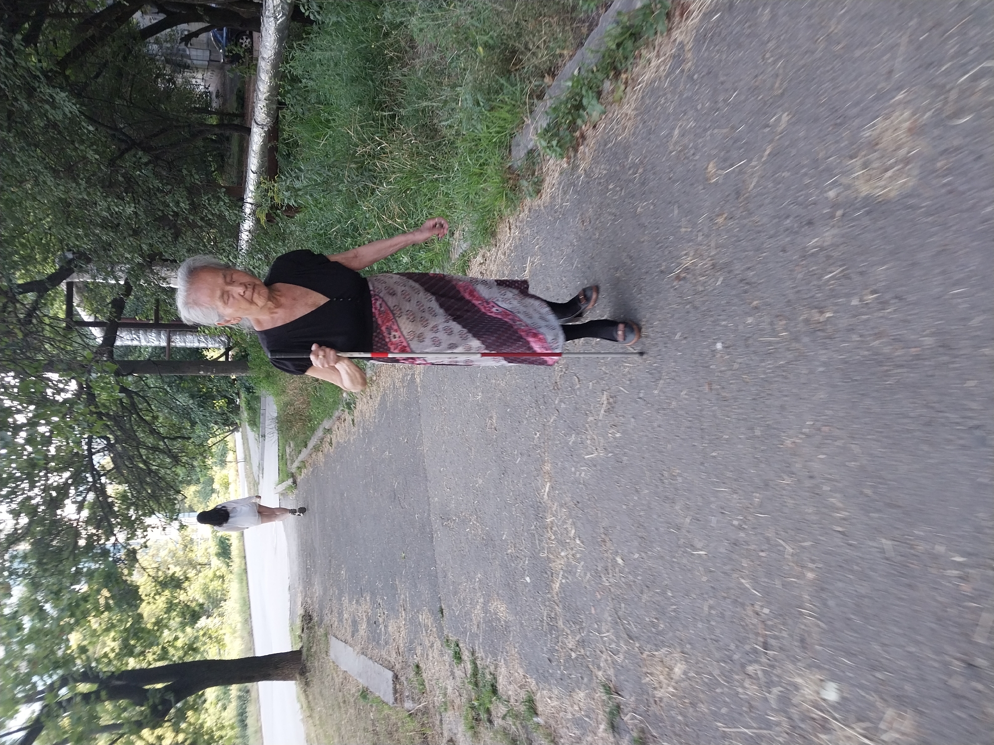 An elderly Asian woman walks on a paved path in Dnipro, Ukraine, with a white cane. She is wearing a black V-neck top and a patterned skirt. A younger woman in a white top and shorts walks in the background on the same path, further away. The path is lined with trees and overgrown grass. A large pipe covered in insulation runs along the right side of the path.